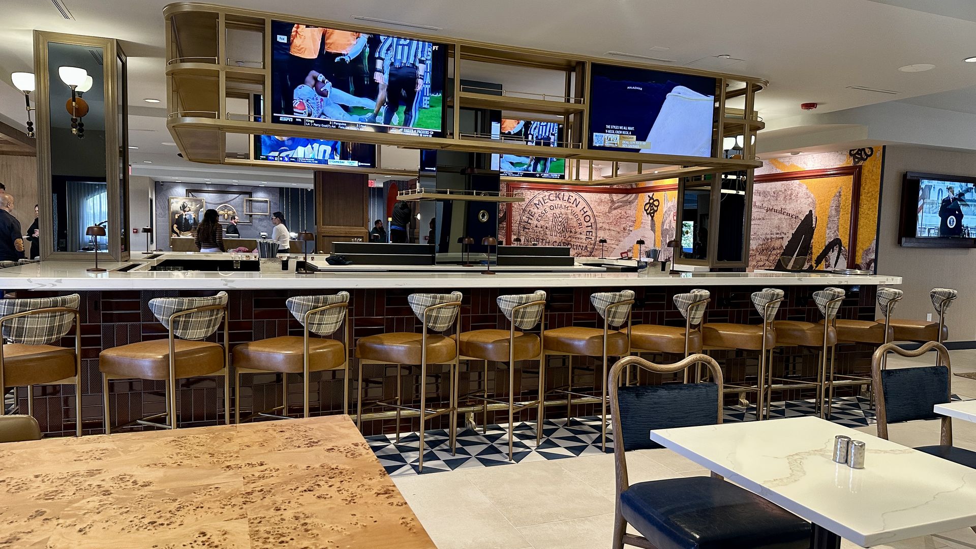 Modern hotel bar with leather stools, white marble countertop, TV screens showing sports, geometric floor tiles, and wooden tables and chairs in a well-lit room.