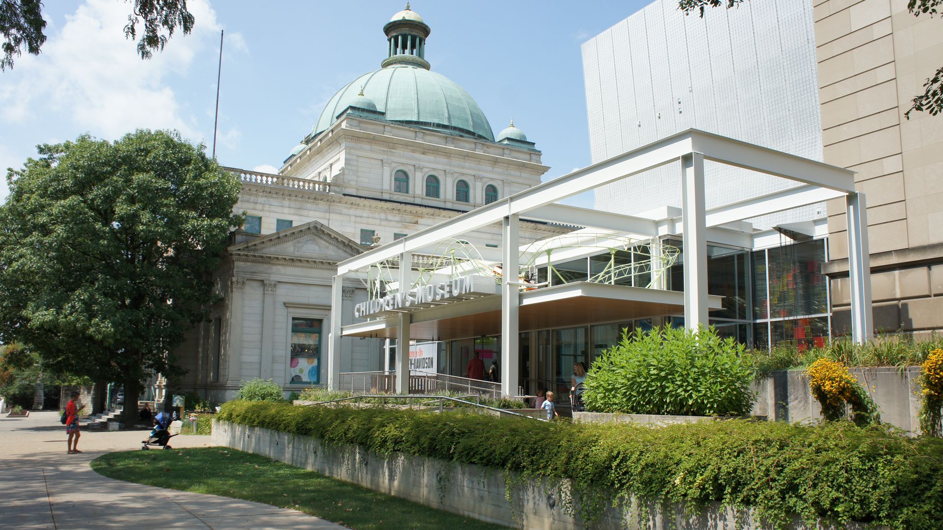 Exterior view of a children's museum with a modern white frame entrance and a large domed historic building in the background, surrounded by greenery and a sidewalk.