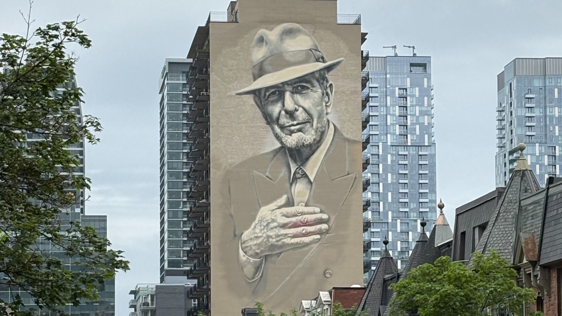 A grayscale mural shows an elderly man in a fedora and suit, hands on his chest, on a tall beige building. Modern glass towers rise behind, with trees in the foreground.