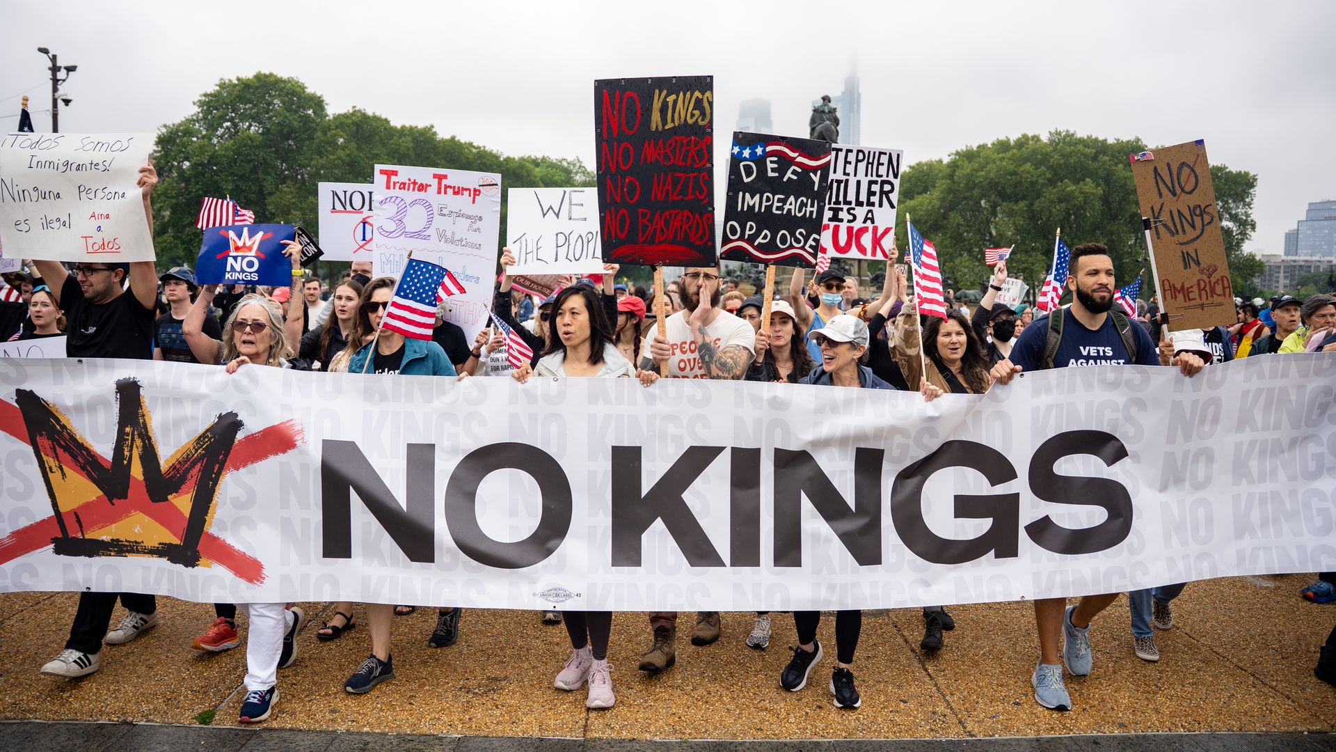 Diverse protesters march in a park, holding a large white banner reading "NO KINGS" with a red crossed-out crown; others display signs and American flags under a cloudy sky.