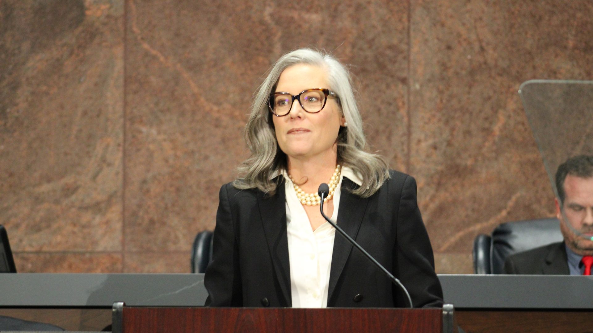 Katie Hobbs stands at a lectern with a microphone in front of her in the Arizona House of Representatives. 