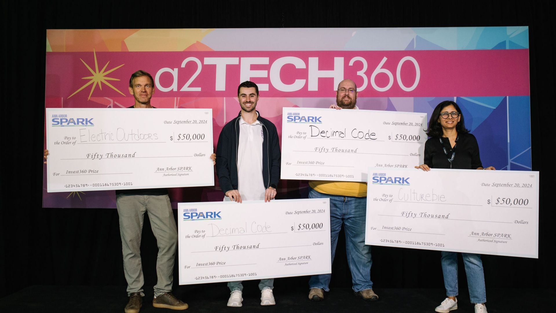 Four people stand in front of a colorful a2TECH360 backdrop holding large $50,000 Ann Arbor SPARK prize checks made out to Electric Outdoors, Decimal Code, and Culturebie.
