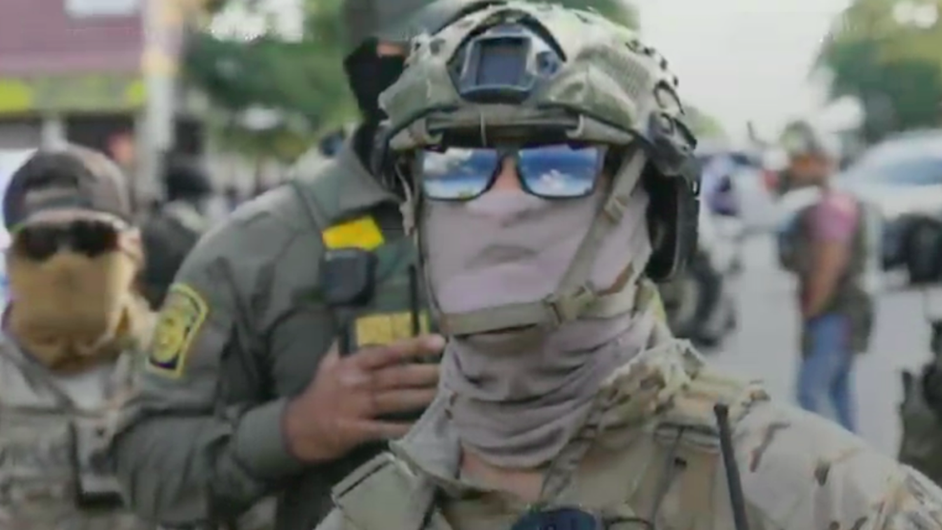 A soldier in desert camouflage wearing a helmet with night-vision gear, scarf over the face, and reflective sunglasses; stands on a street with other troops in a blurred background.