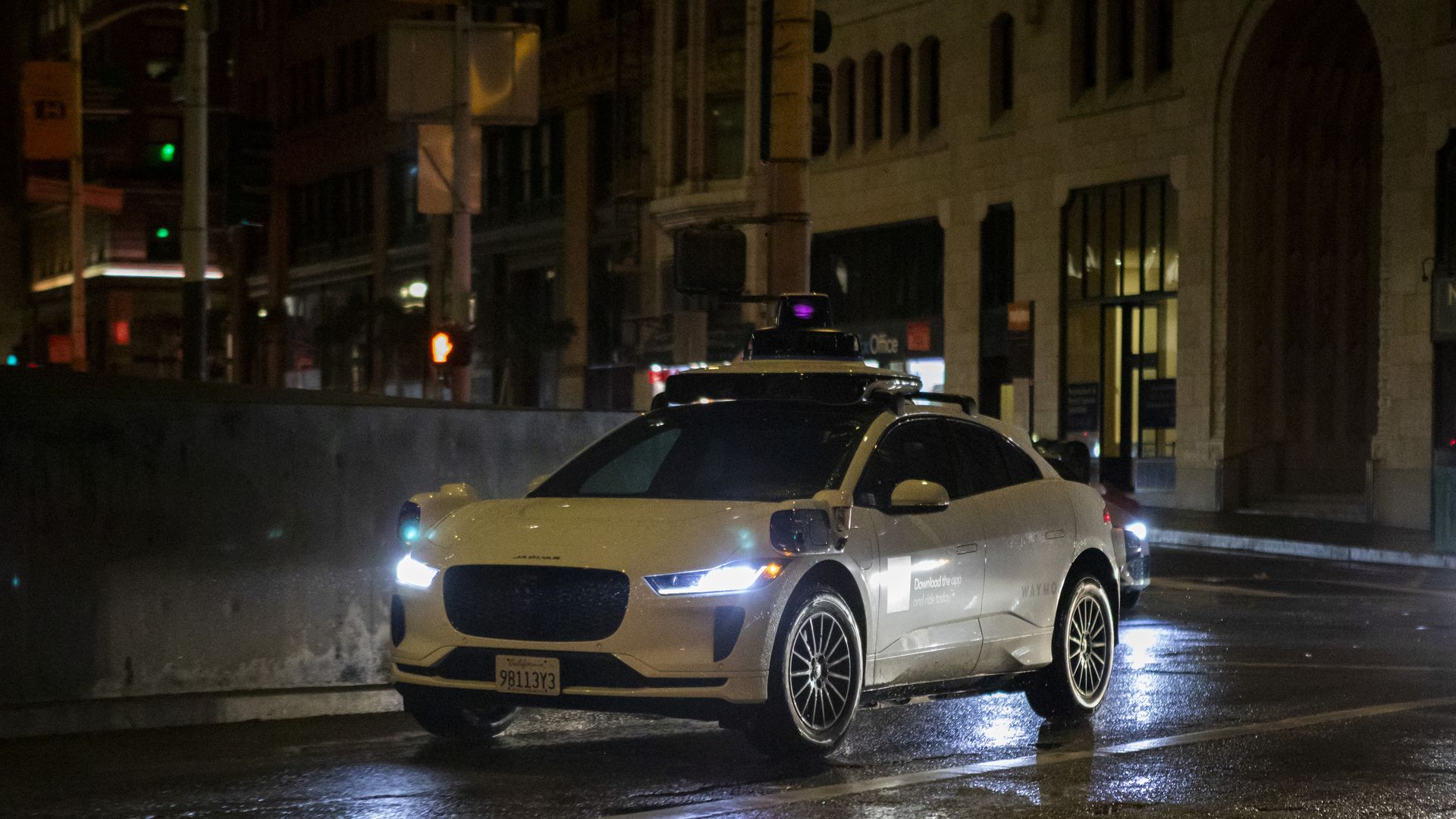 Night city street with a white Waymo robotaxi in the foreground. Wet pavement reflects streetlights as tall buildings loom in the background.