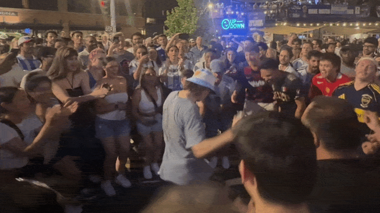 Nighttime crowd forms a circle around a man in a light blue shirt and hat dancing center; many spectators film with phones as a neon blue sign reading "UP-DOWN" glows in the background.