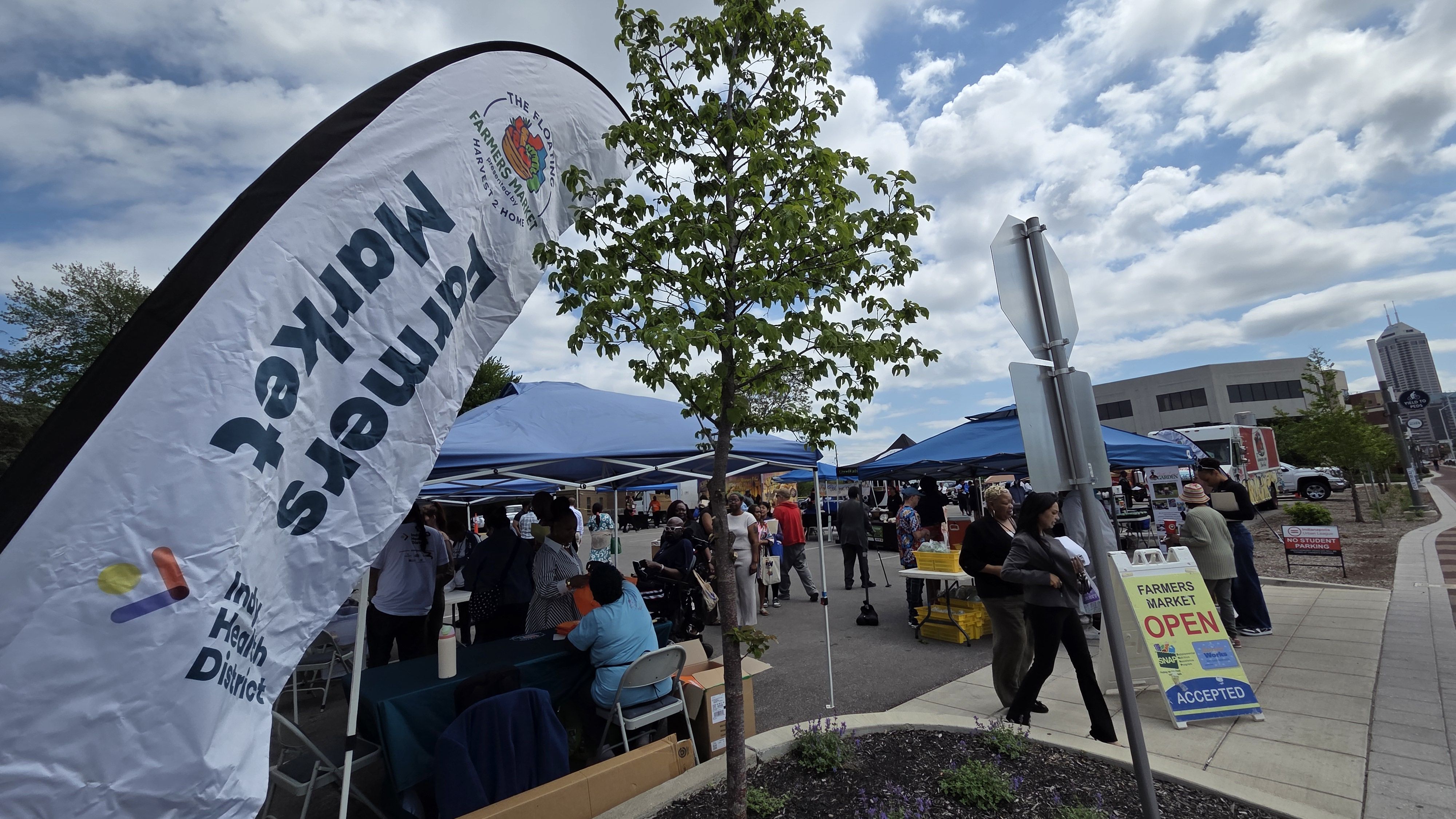 Outdoor farmers market under a blue sky with clouds. White banner and blue tents line the walkway as people browse stalls; a FARMERS MARKET OPEN sign sits near a tree with buildings in the background.
