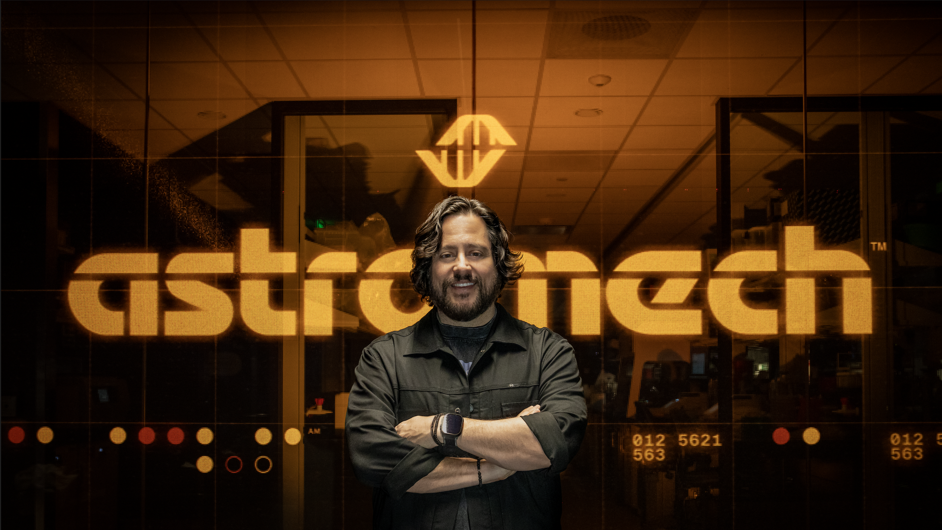 A bearded man with wavy hair in a dark jacket stands with arms crossed in front of an orange-tinted glass wall featuring a large glowing logo and bold letters in the background.