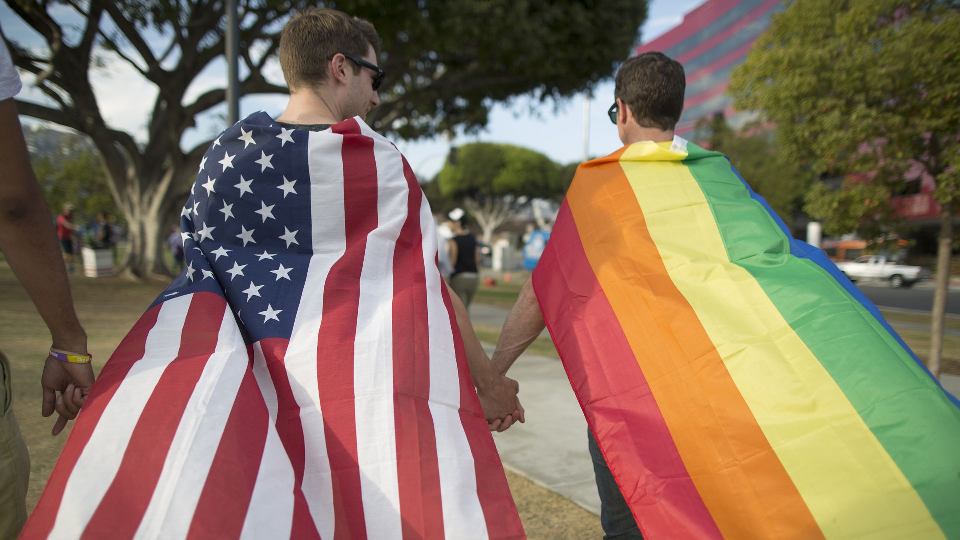 Two men, one wearing an American flag and one wearing a LGBTQ rainbow flag, hold hands.