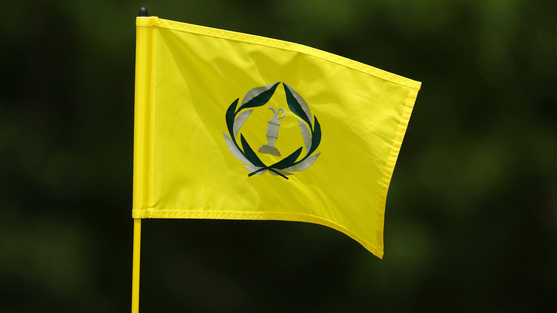 Close-up of a bright yellow Memorial Tournament pin flag on a pole, featuring a green laurel wreath around a silver torch emblem, with a dark green, out-of-focus background.