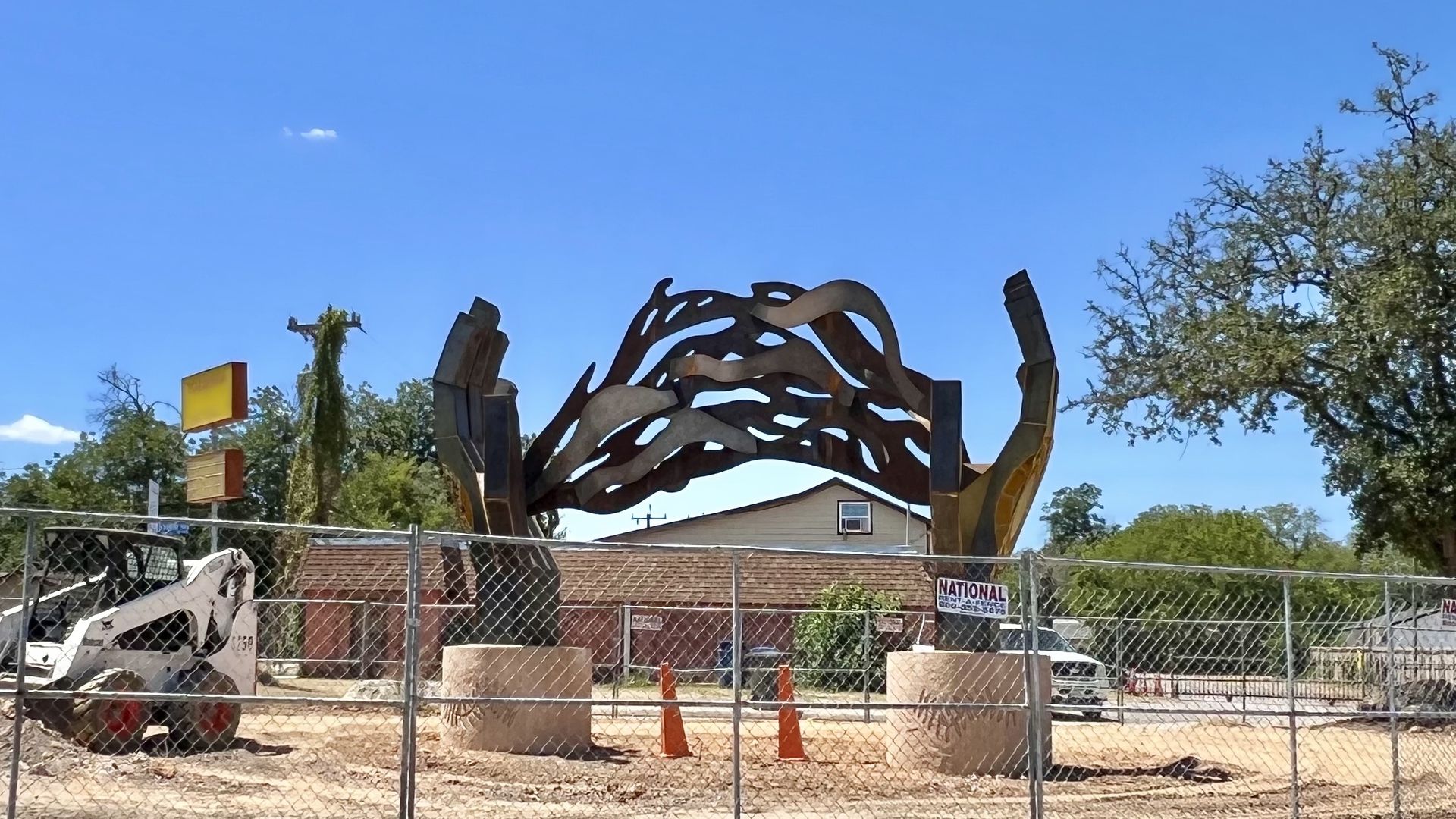 Metal sculpture resembling hands forming an arch with abstract shapes between them, surrounded by a chain-link fence and construction equipment under a clear blue sky.