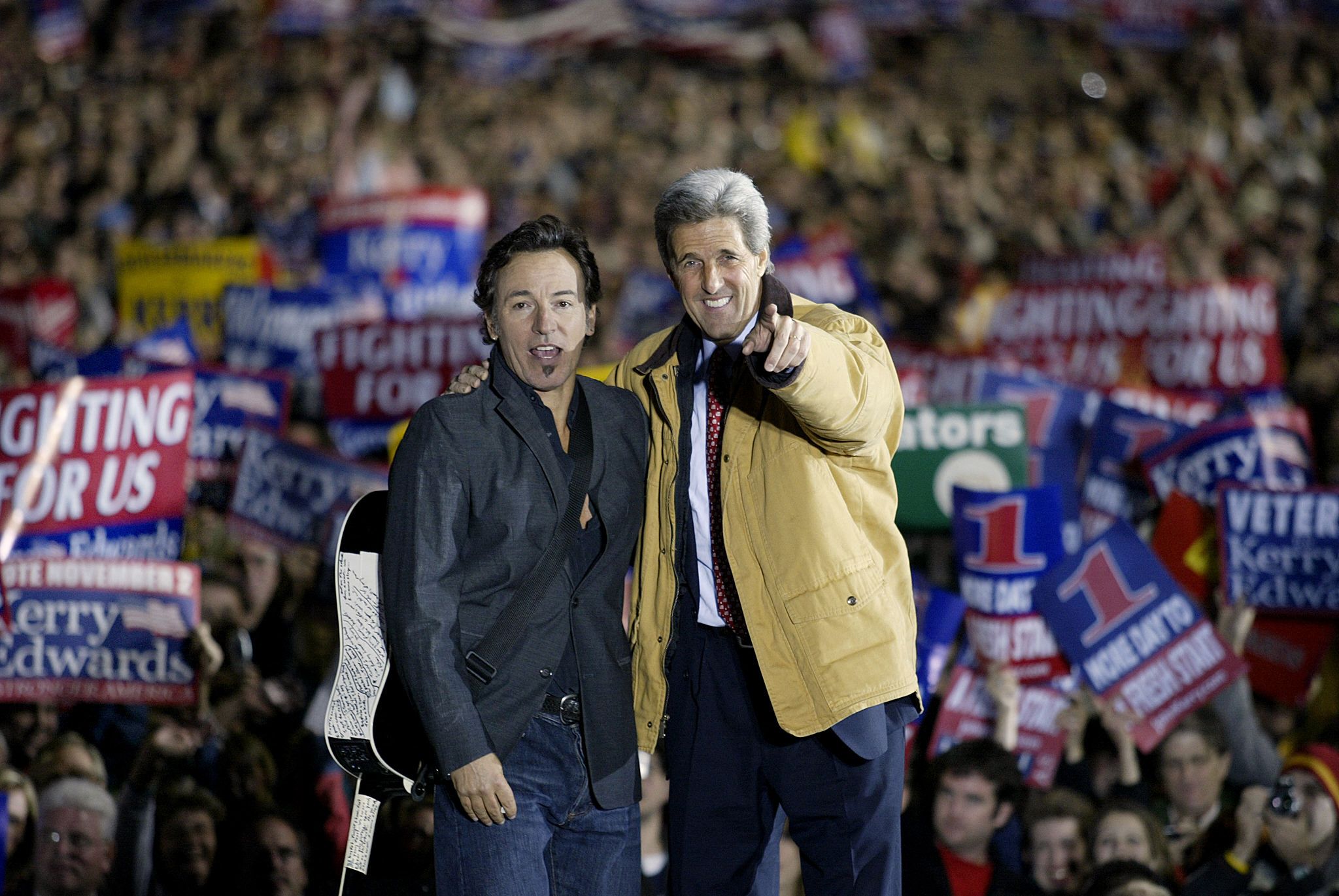 Two men stand on a stage at a crowded political rally; one in a dark suit with a guitar, the other in a tan coat points at the camera. Behind them, a sea of supporters holds red, white and blue signs.