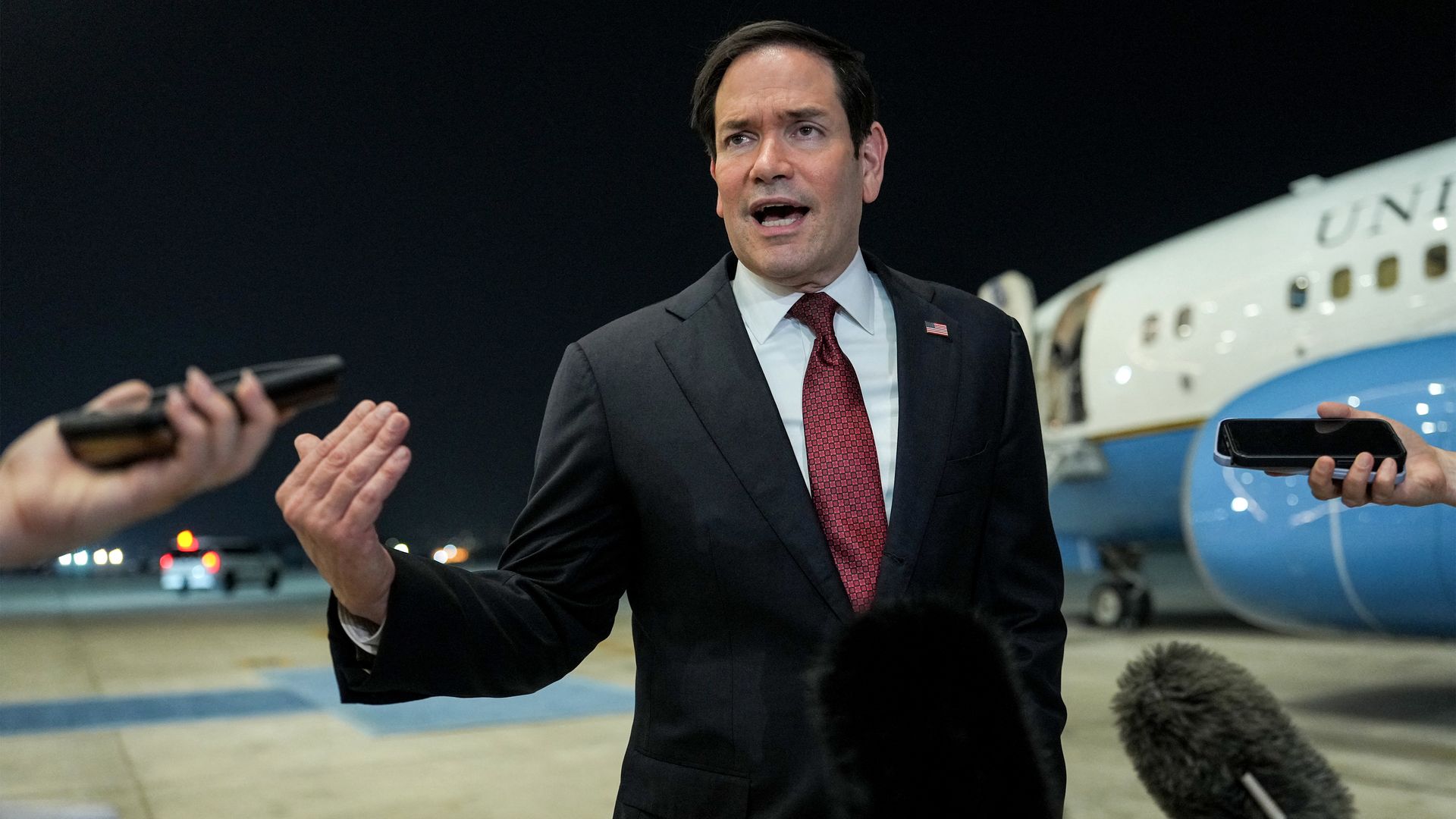 A man in a dark suit and red tie speaks to reporters on an airport tarmac at night; a white and blue United Airlines plane sits behind him as microphones are held up nearby.