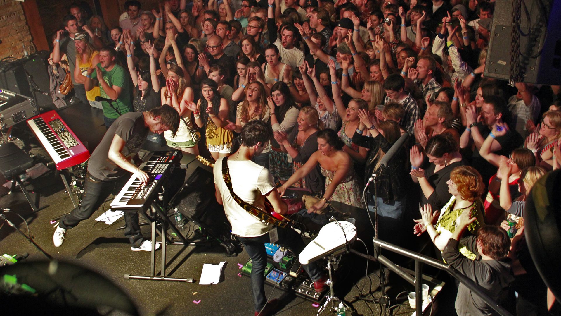 A band on the stage at a music venue as concert-goers cheer, many with their hands raised