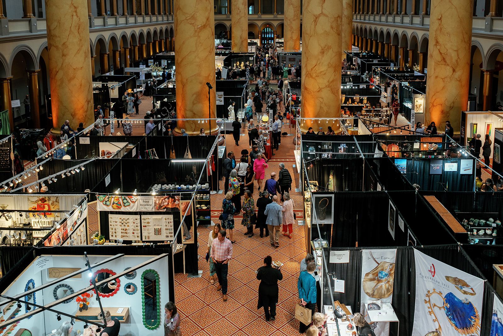 A grand indoor expo hall with tall marble columns, rows of black exhibition booths, and a crowd browsing jewelry and crafts along a patterned tile floor.