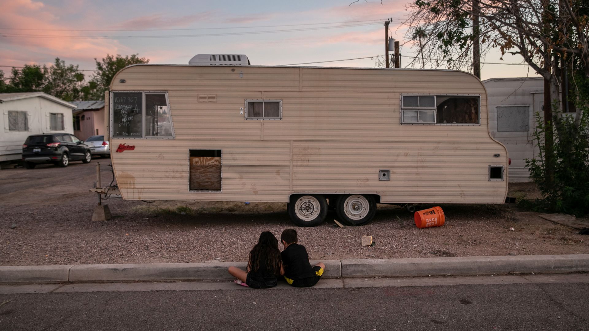 Two children sit in front of an RV on the curb 