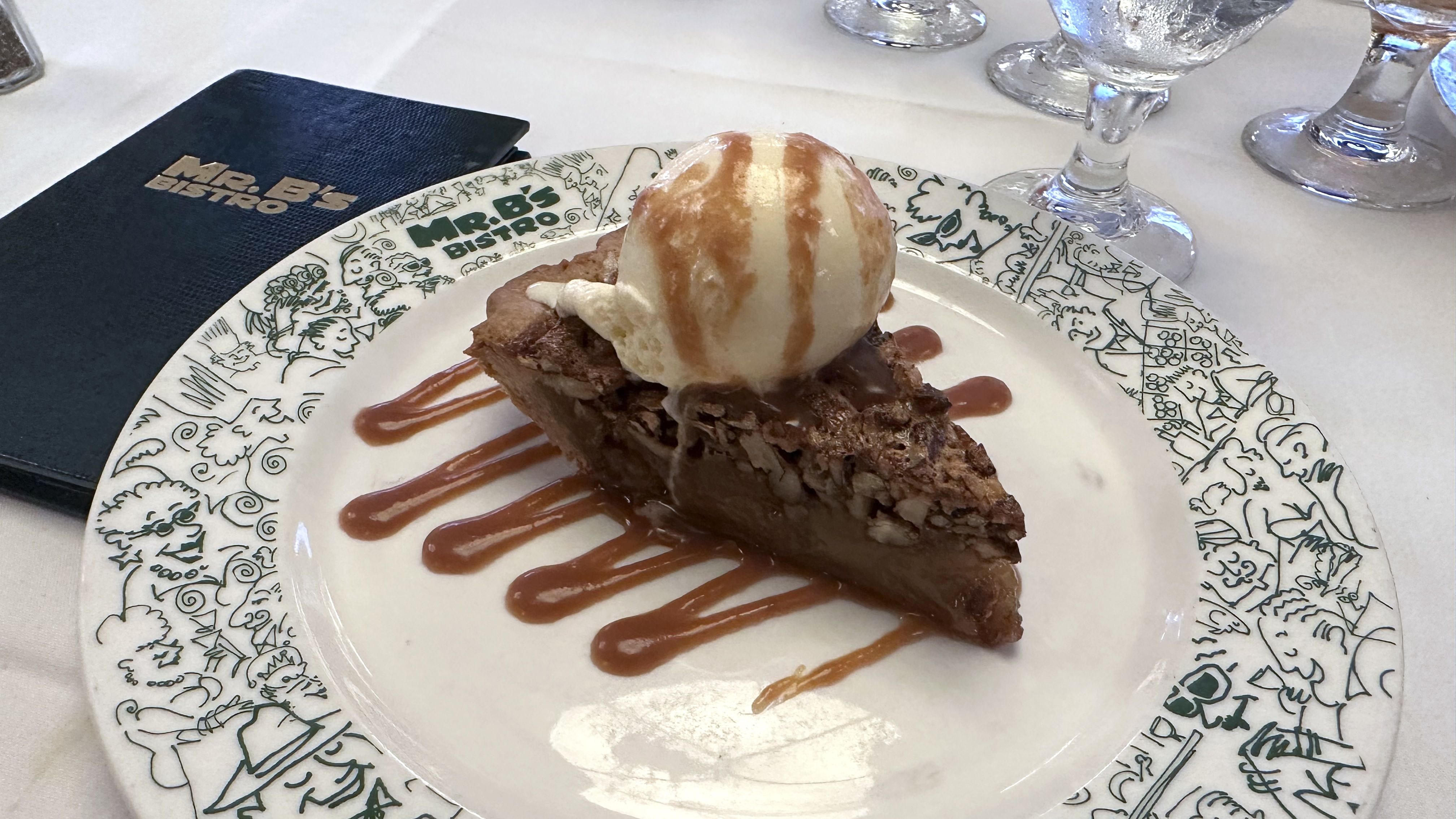 Slice of pecan pie topped with a scoop of vanilla ice cream and caramel drizzle on a white plate with green cartoon designs and "Mr. B's Bistro" logo, next to a black menu.