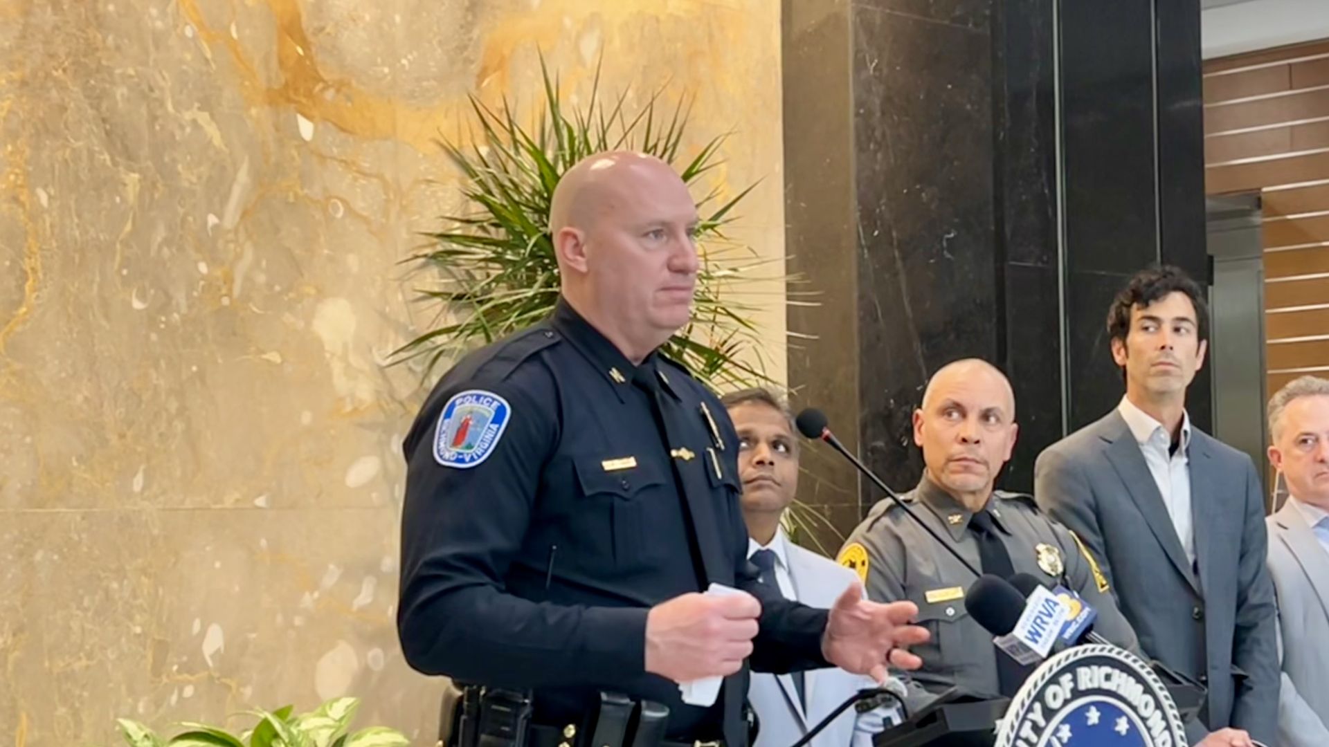 Police officer speaking at a podium with a City of Richmond seal, accompanied by three men in suits and uniform, against a marble wall and green plant background.