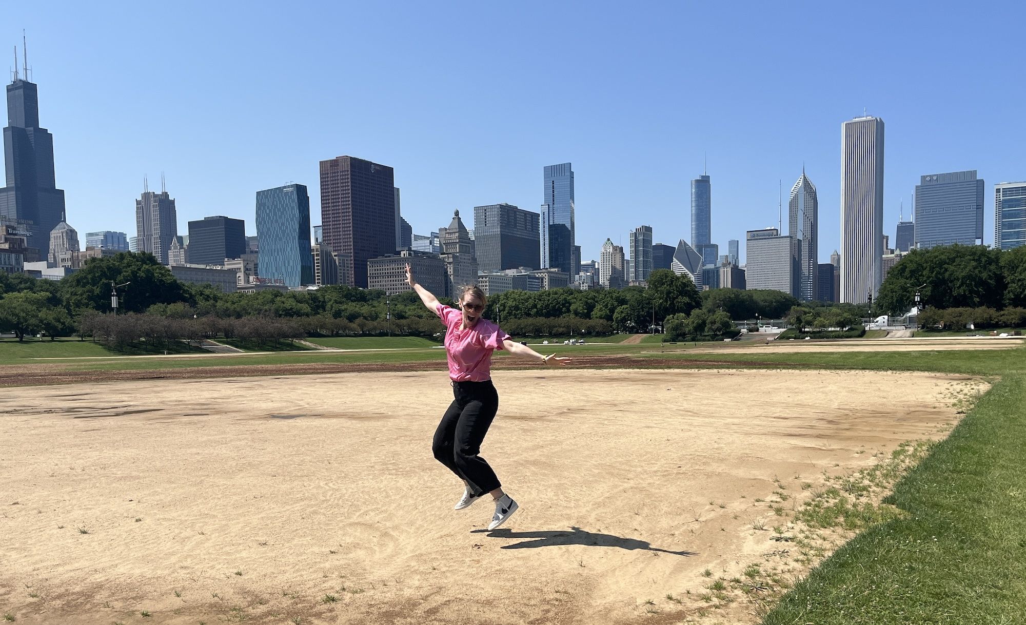 Woman in pink shirt and black pants jumping in the middle of a baseball diamond with the Chicago skyline in the back.