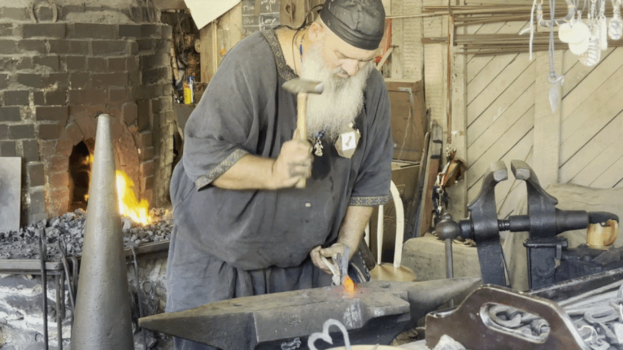 Bearded blacksmith in a dark tunic hammers hot metal on an anvil in a rustic workshop; a glowing forge, a large vise, and various tools hang on the walls.
