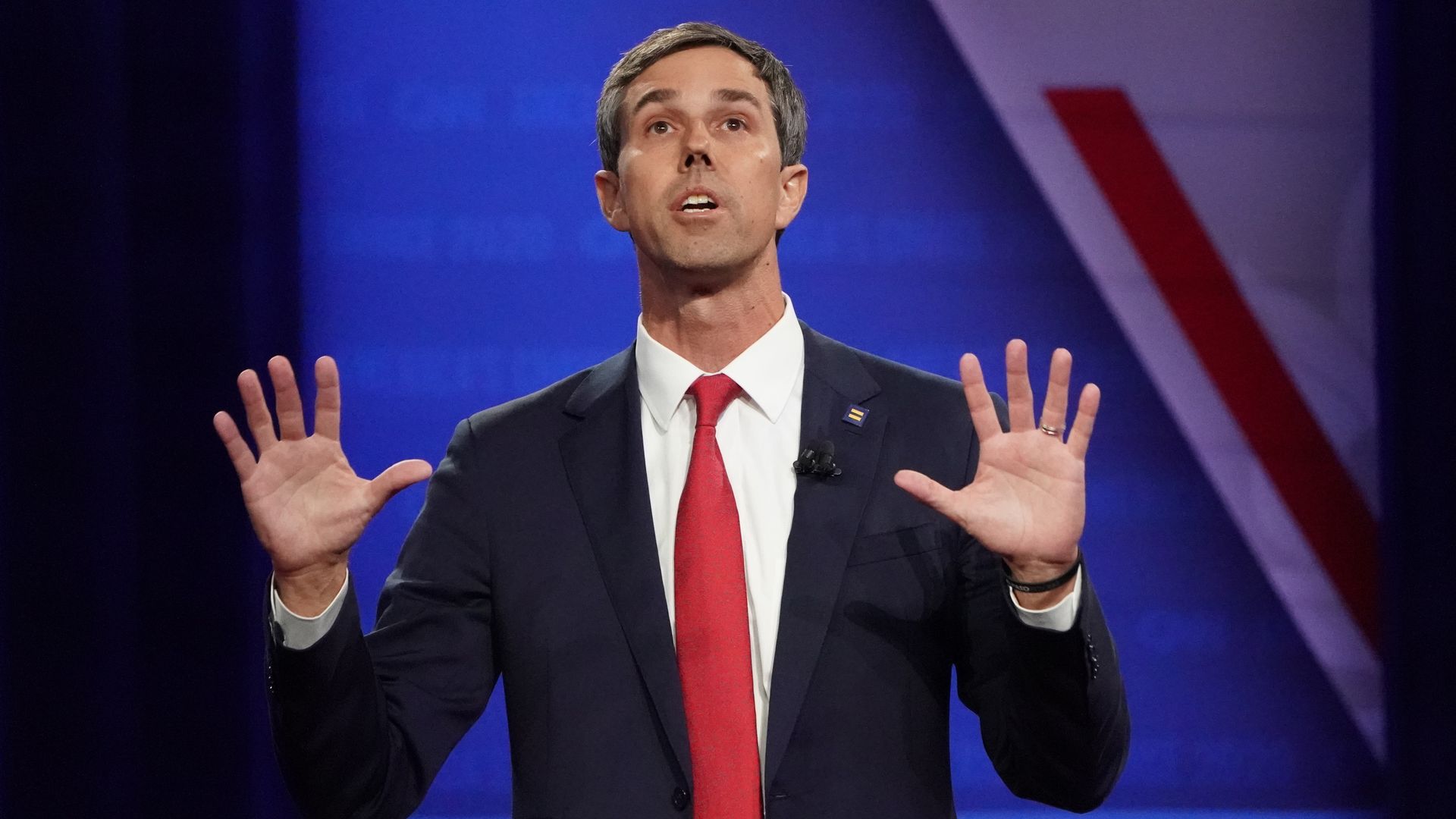 In this image, O'Rourke stands with both hands raised and palms outstretched as he stands on stage in a suit and talks.