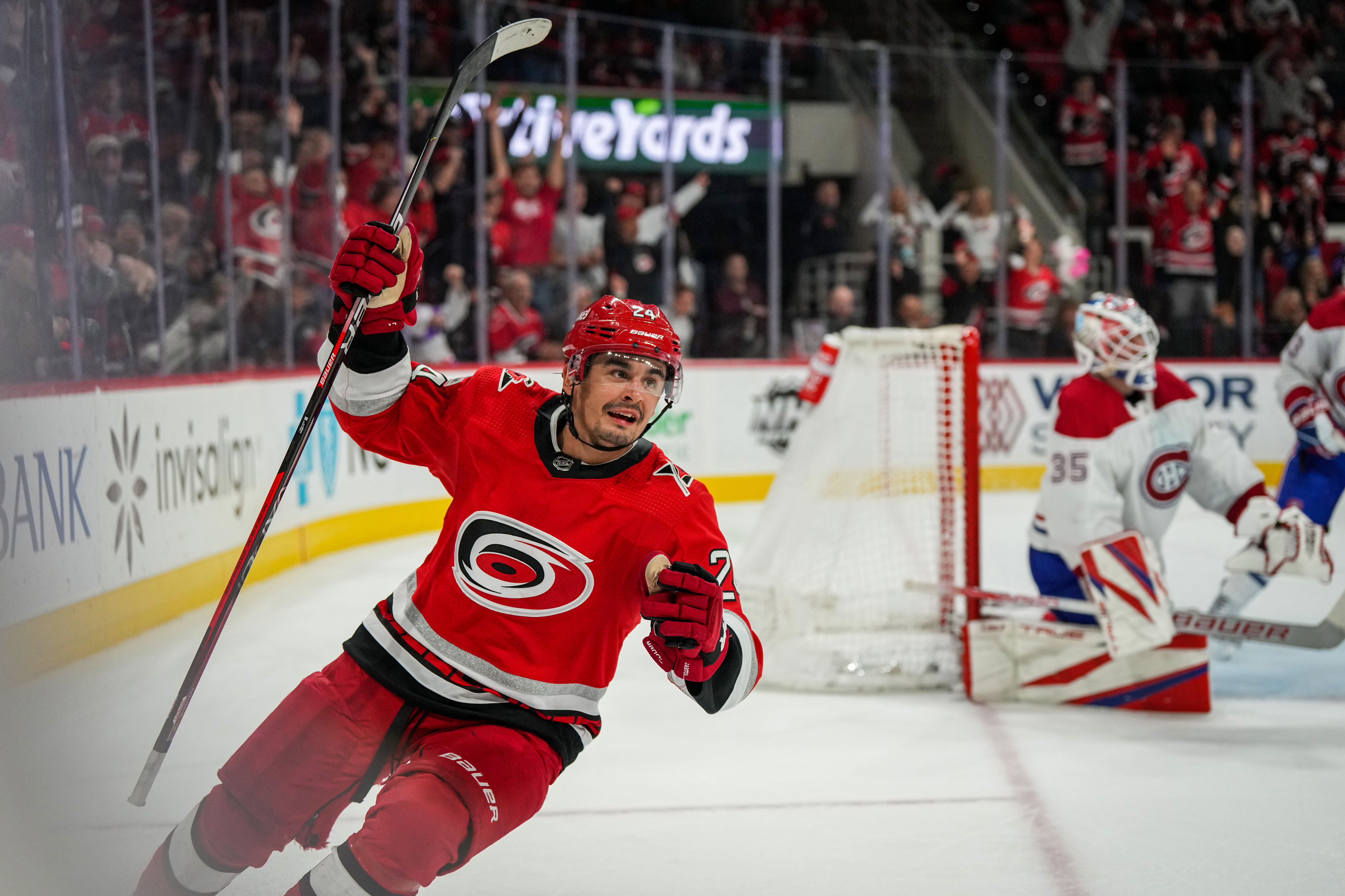 A Carolina Hurricanes player wearing a red jersey raises his stick in celebration after scoring a goal against the Montreal Canadiens, with the opposing goalie and crowd in the background.