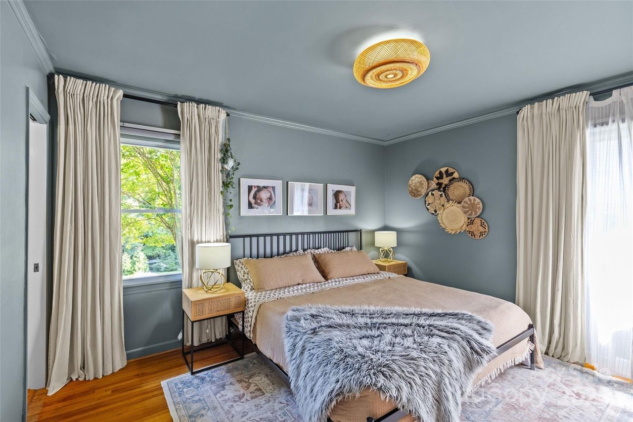 Bedroom with blue-gray walls, beige bedspread, fluffy gray throw, wooden floors, cream curtains, two nightstands with lamps, framed photos above bed, round woven wall decor, and a ceiling light.
