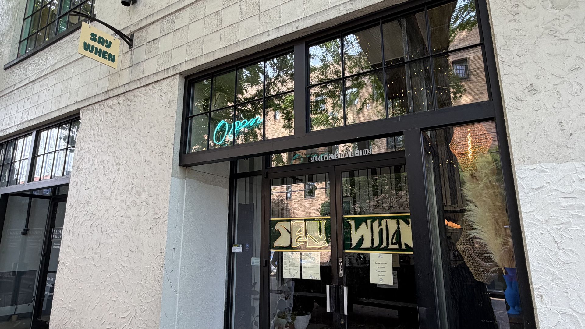 Storefront with a beige textured wall, a hanging sign reading SAY WHEN, large glass doors, turquoise neon Open sign, and reflections of trees and brick buildings.