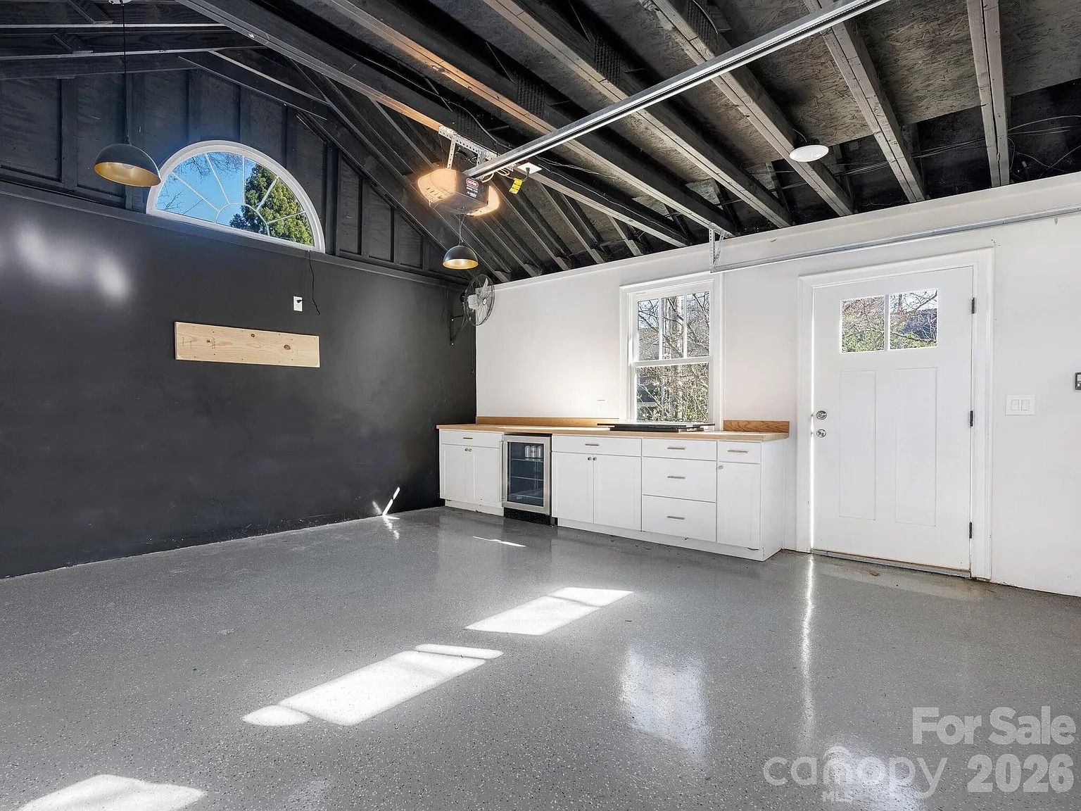 Empty garage with polished gray floor, black wall with arched window, white cabinets with wooden countertop, white door with window, exposed dark wooden ceiling beams, and hanging lights.