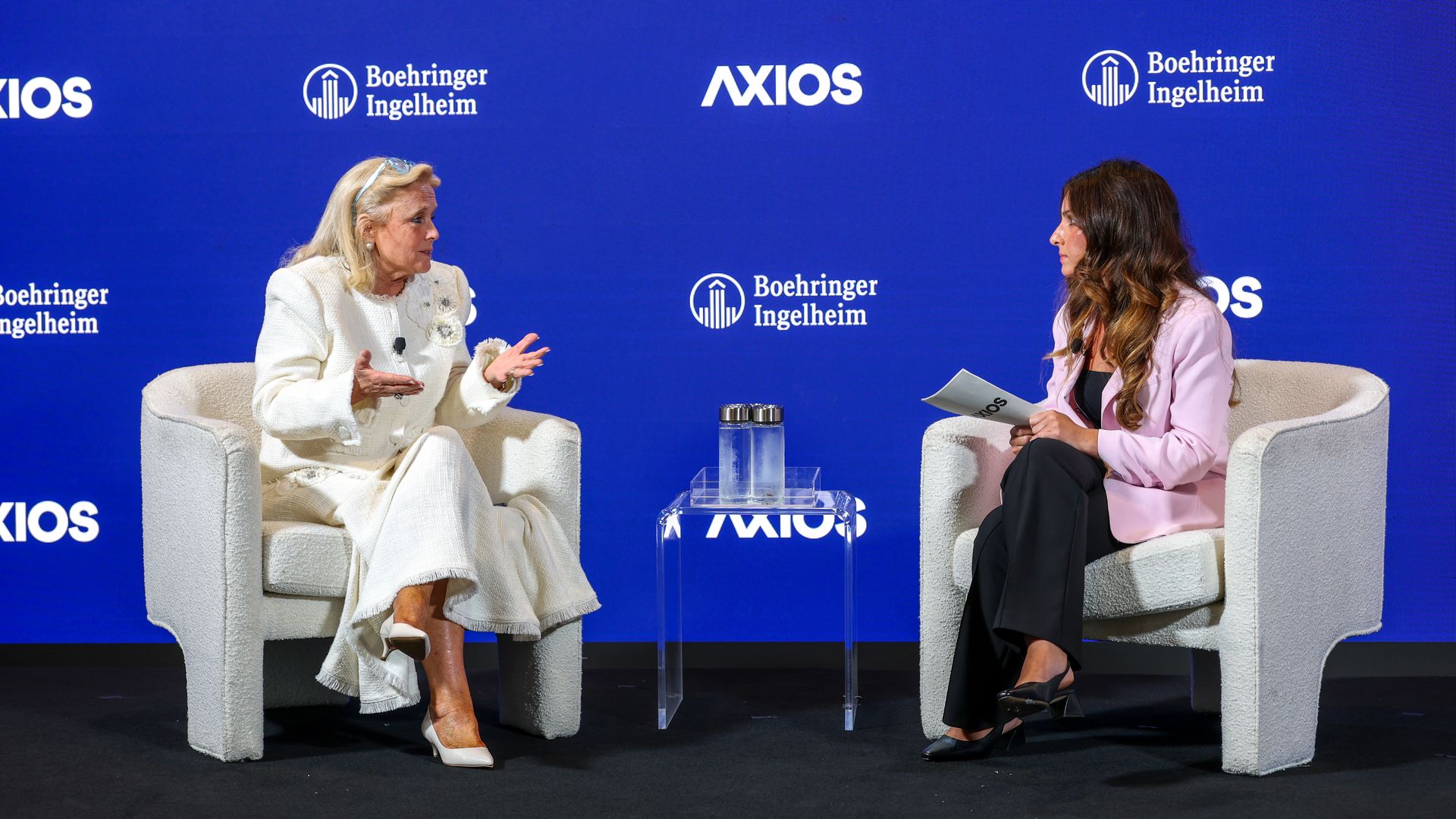 Two women sit in armchairs on a stage, one in white and the other in pink, discussing with microphones; a blue backdrop shows Axios and Boehringer Ingelheim logos, with a clear glass table and water bottles between them.