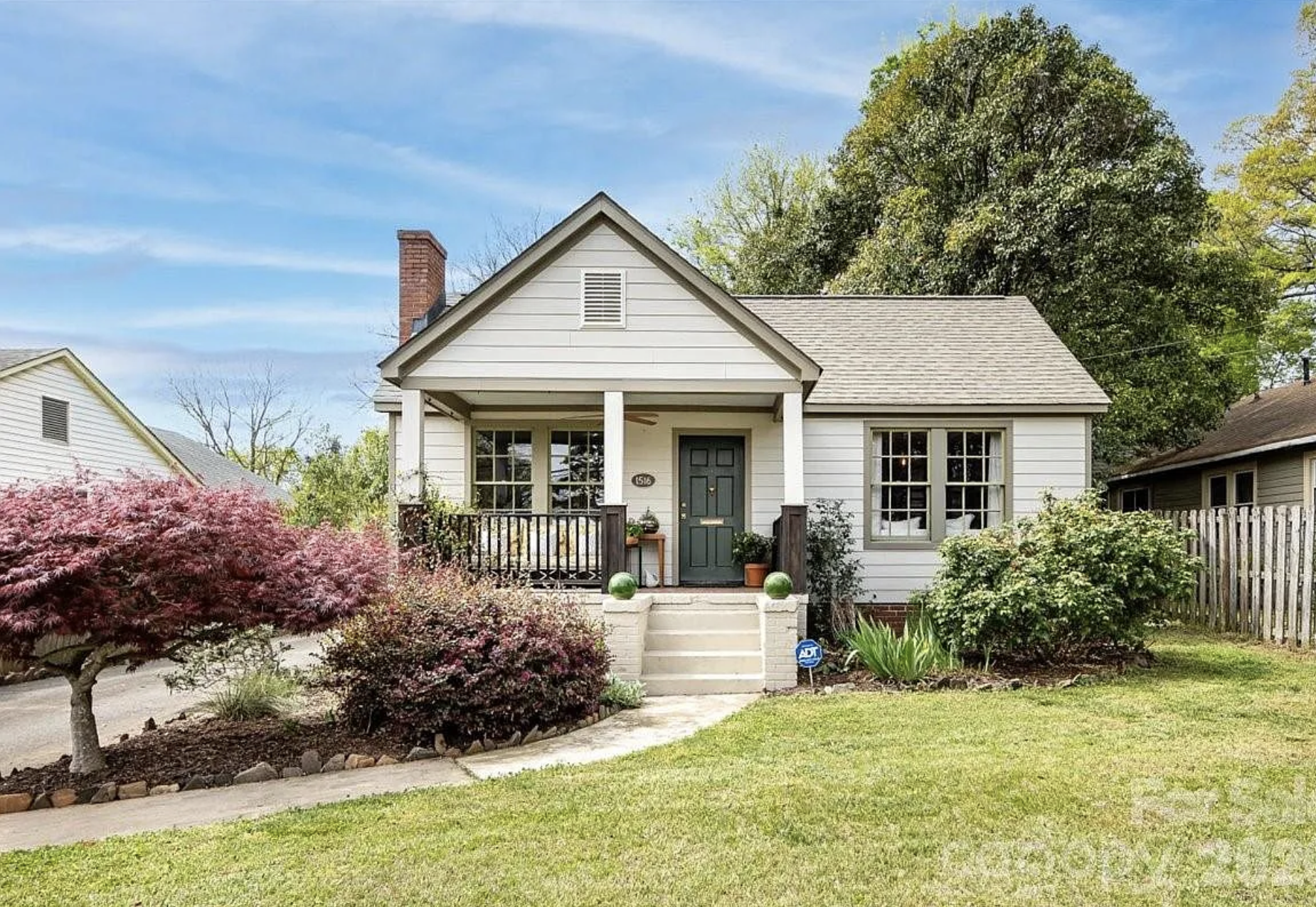 Cozy, single-story white cottage with a gray gabled roof, green front door, and small porch. Red-leaf shrub left, green bushes right, blue sky, brick chimney, and a fenced yard.