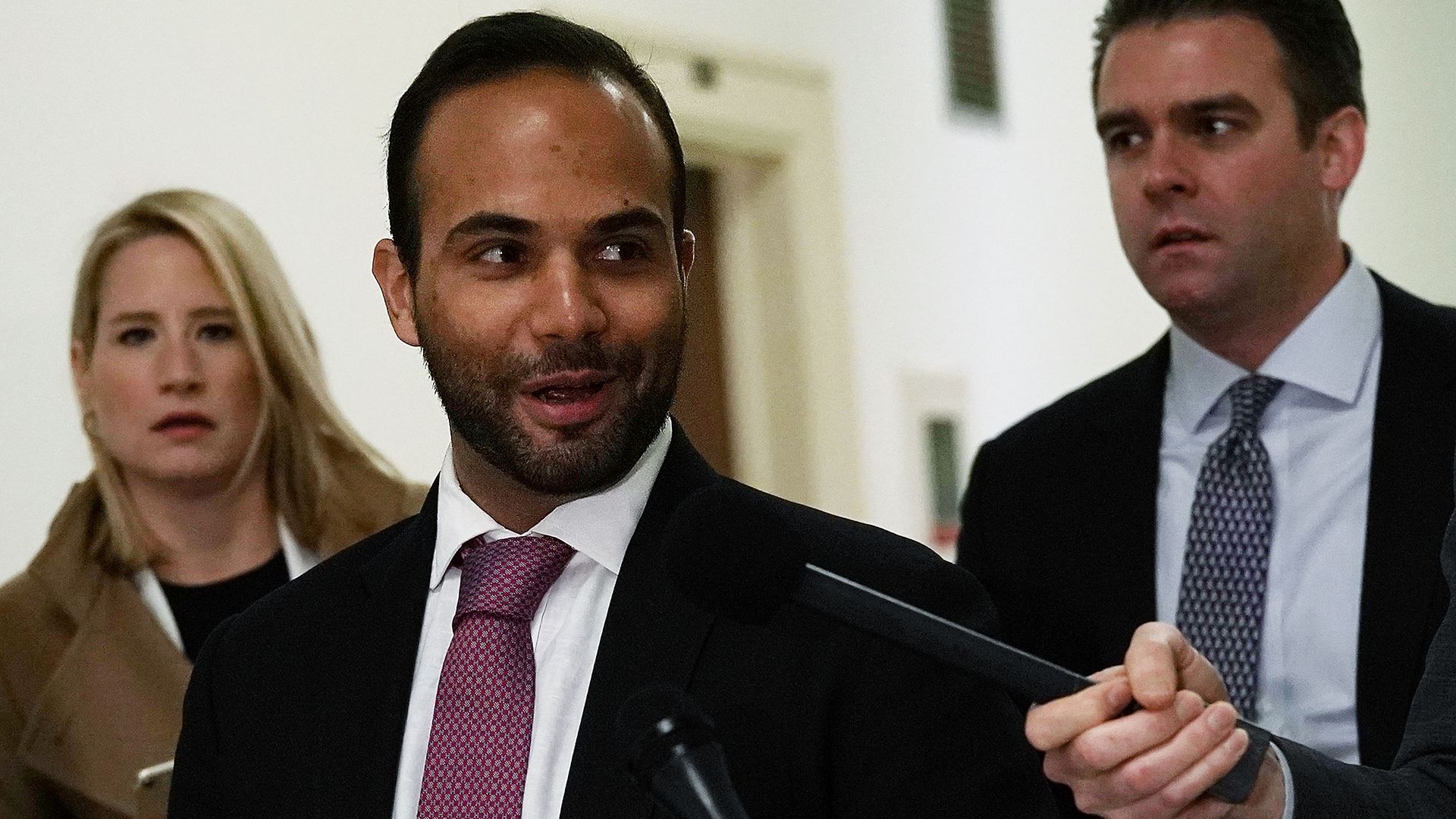 In this image, George smirks and speaks into a microphone while wearing a suit.