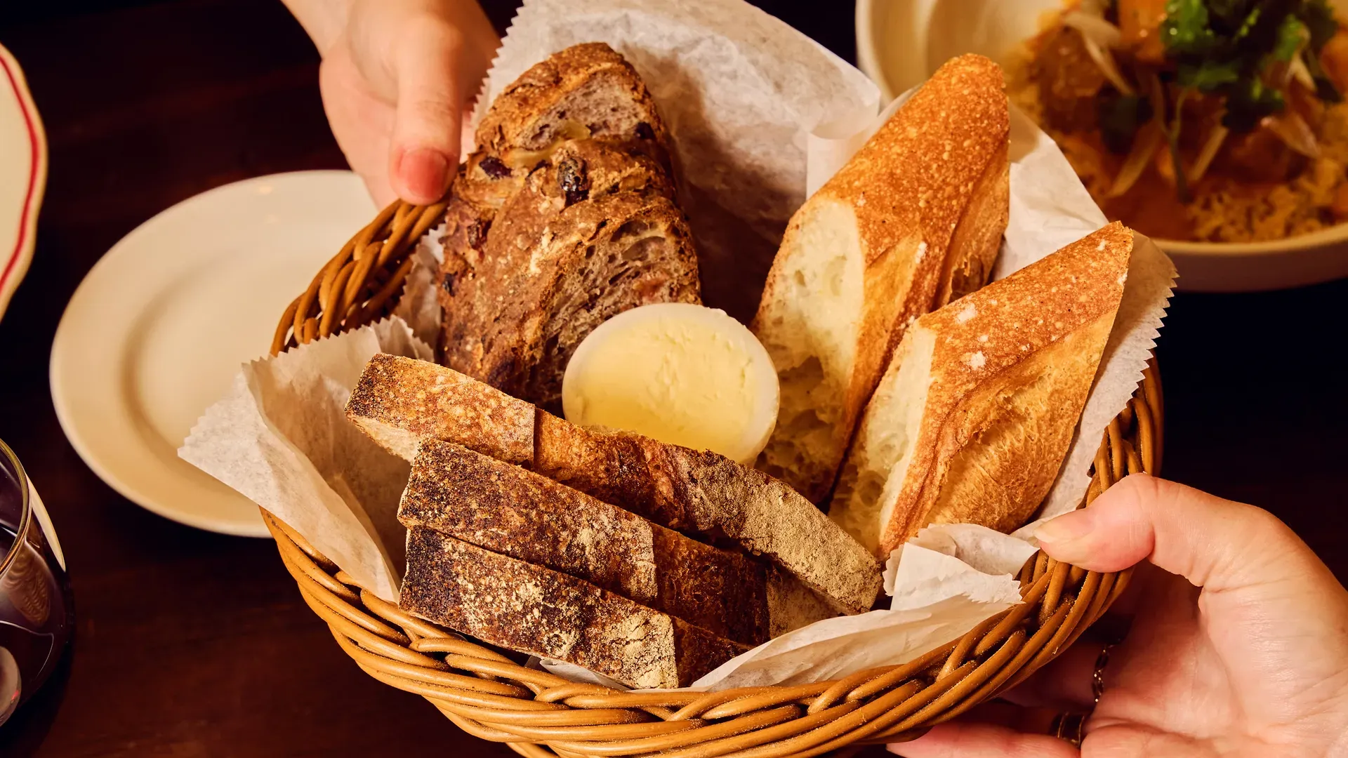 A wicker bread basket lined with parchment holds an assortment of baguette and rustic loaves, a pat of butter in the center, and hands steadying the basket in a dining setting.