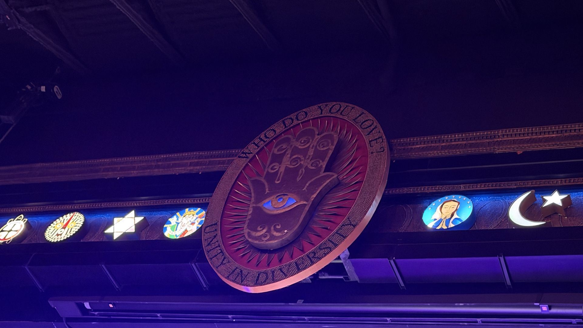 Dimly lit venue ceiling with a large circular medallion featuring an eye at the center and text around the rim. Below, a row of circular icons including stars and a crescent moon.