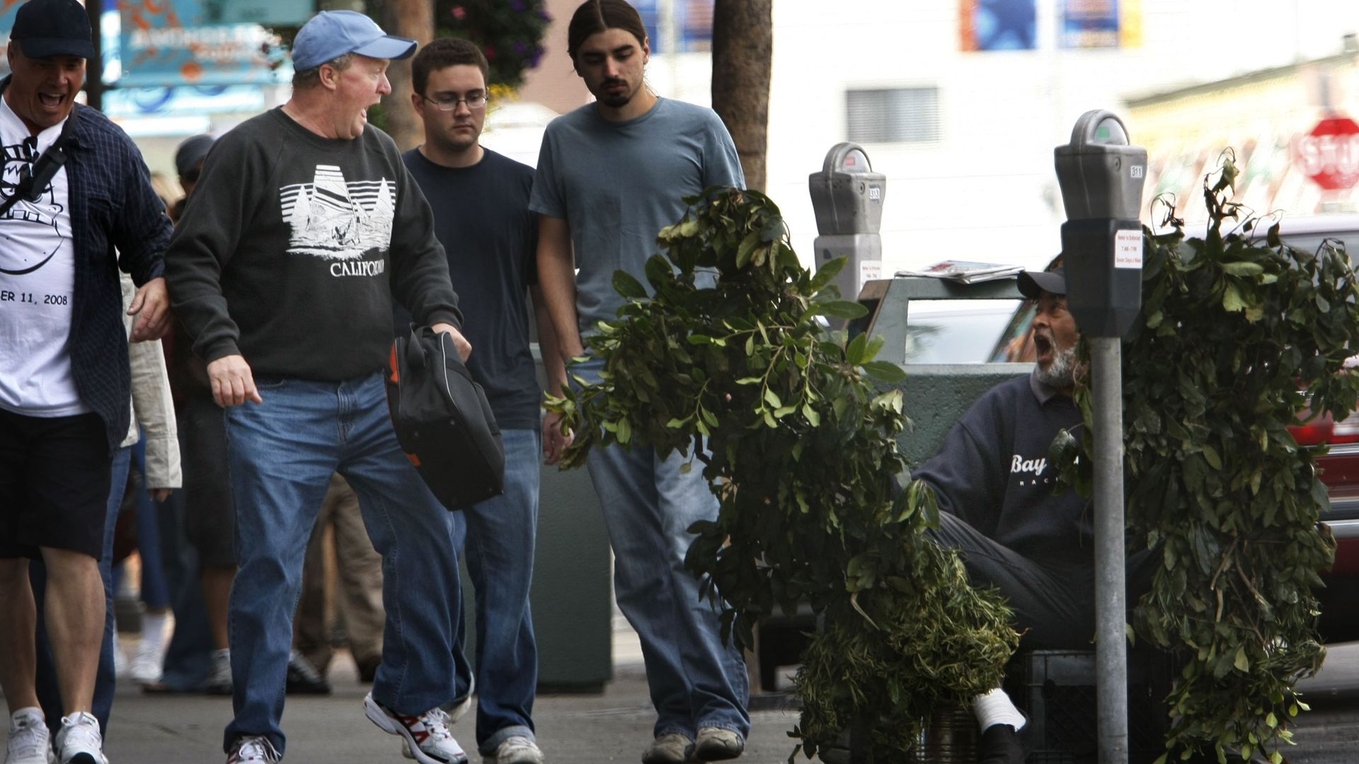Man walking down street next to man hiding behind bush.