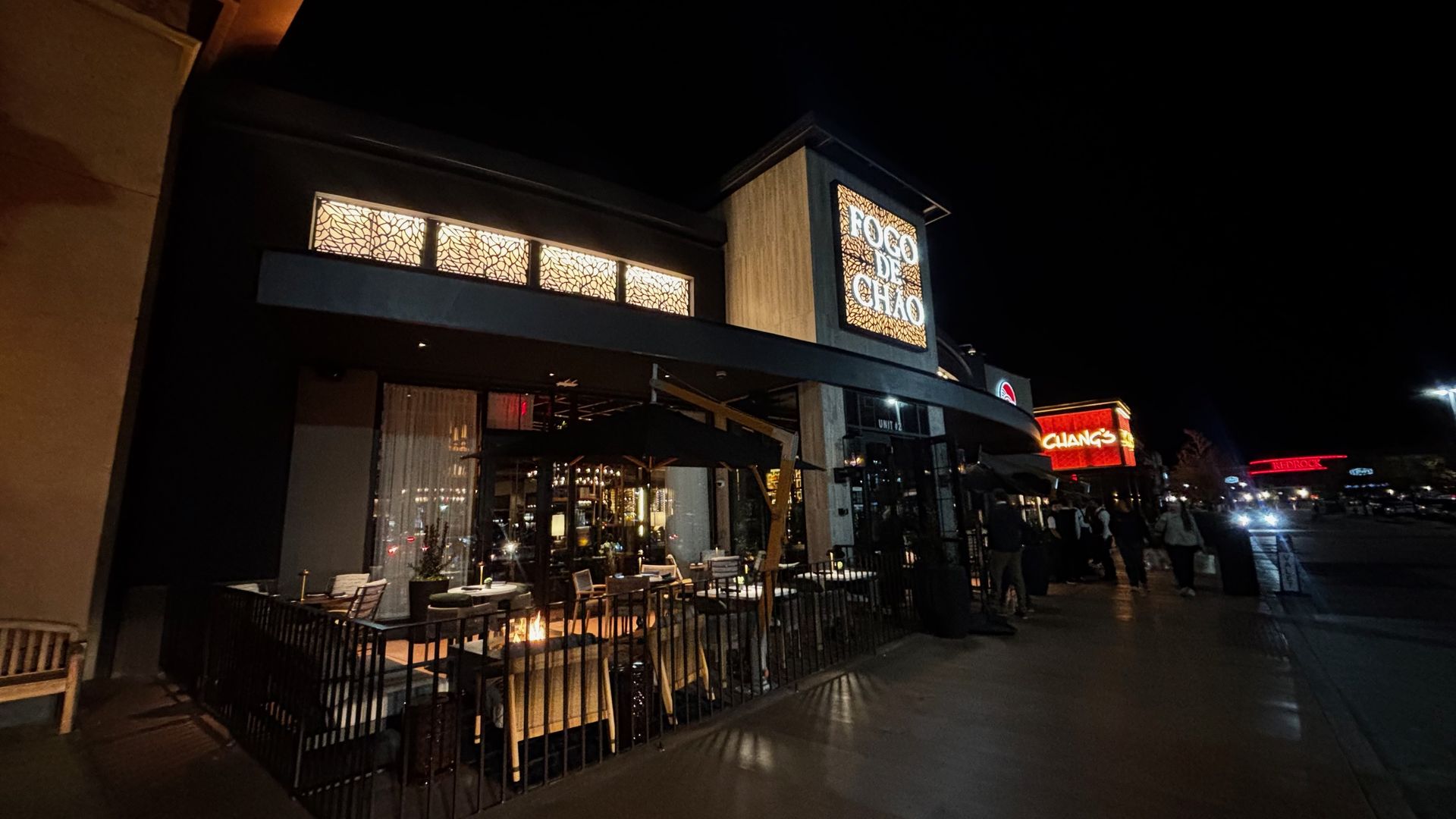 Night scene of a restaurant exterior with warmly lit patio seating and a glowing sign reading "FOGO DE CHÃO", alongside other lit business signs in the background.