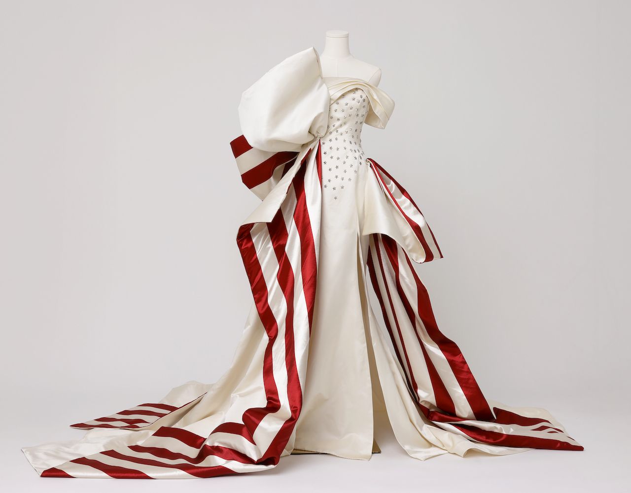 Ivory couture gown on a mannequin with an off-the-shoulder bodice, star embellishments, and a dramatic oversized bow. A bold red-and-ivory striped train trails behind.