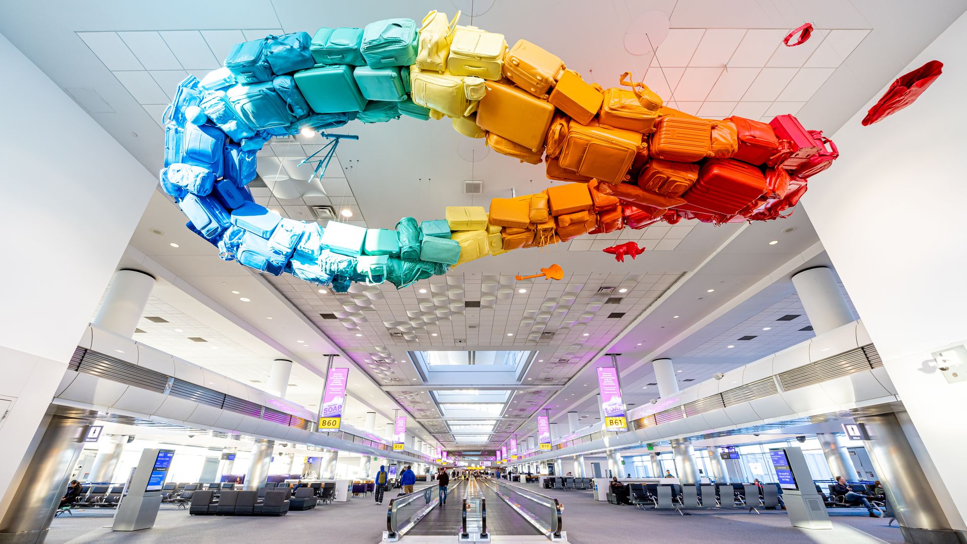 A large, colorful suspended art piece made up of luggage bags hangs over an airport concourse. 