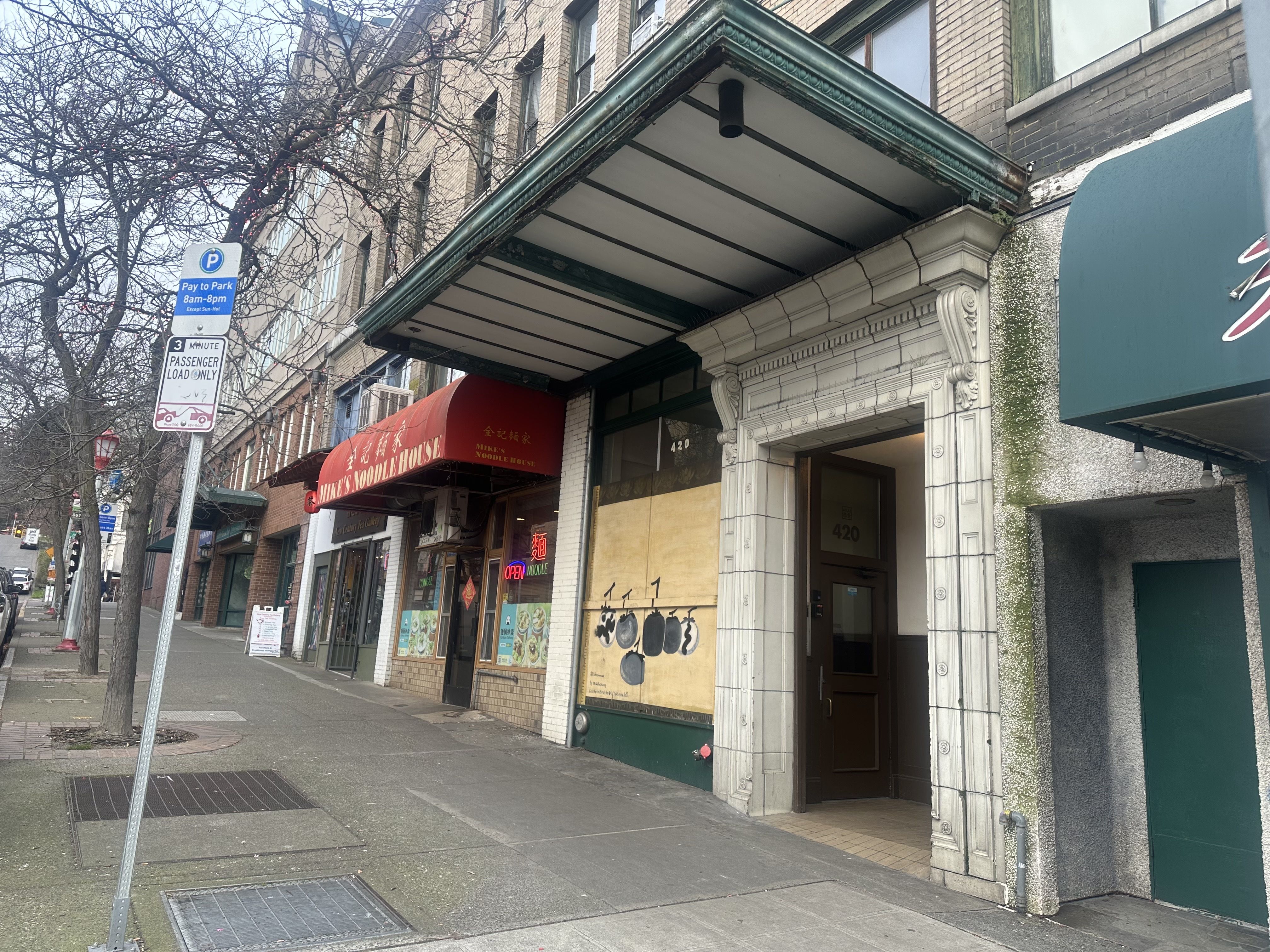 Street view of a sidewalk with leafless trees, a pay-to-park sign, and storefronts including Mine's Noodle House with a red awning and an entrance marked 420 with decorative stonework.