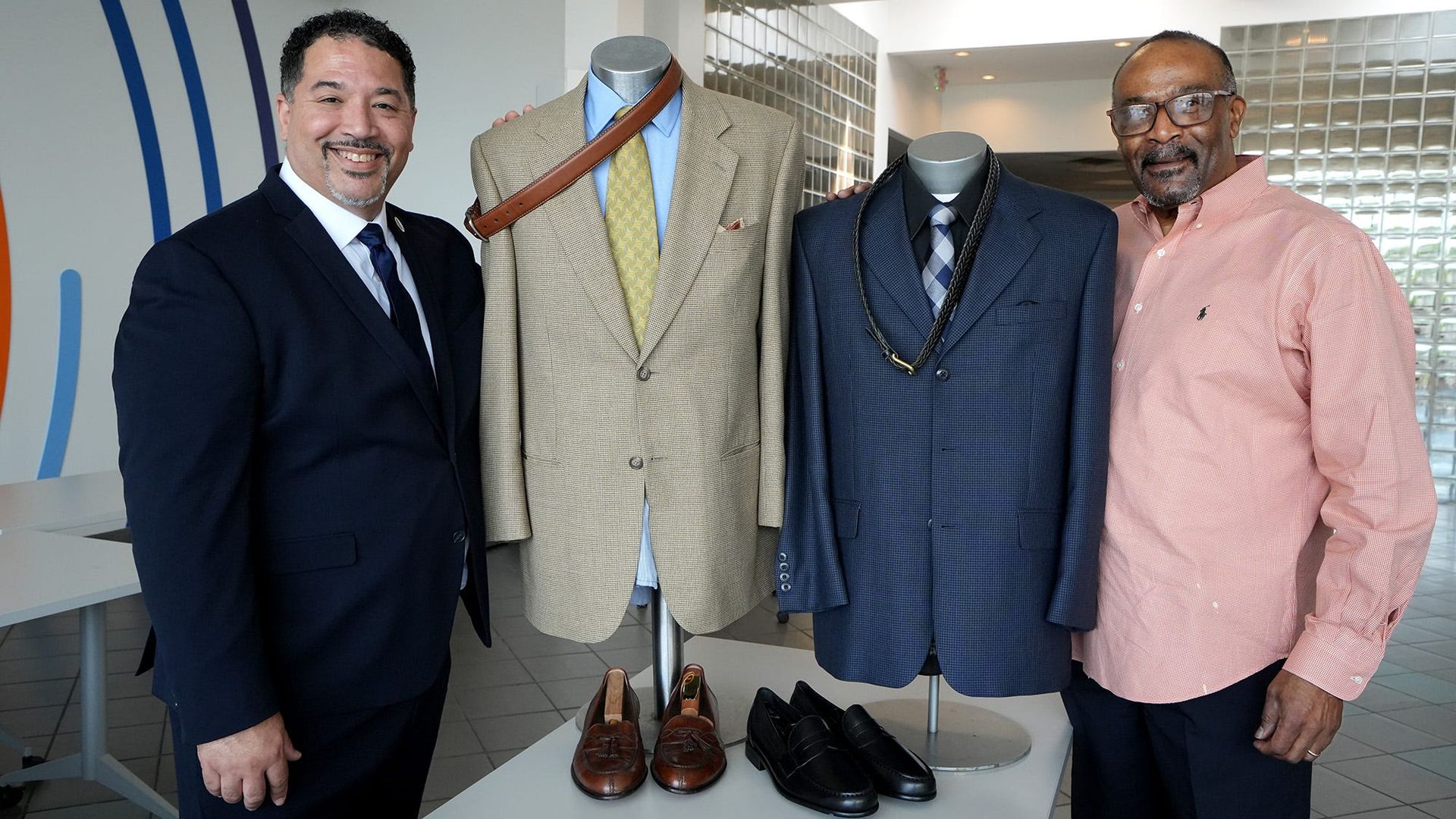 Two men stand beside suit displays at a nonprofit offering low-income men with professional warddrobes. 