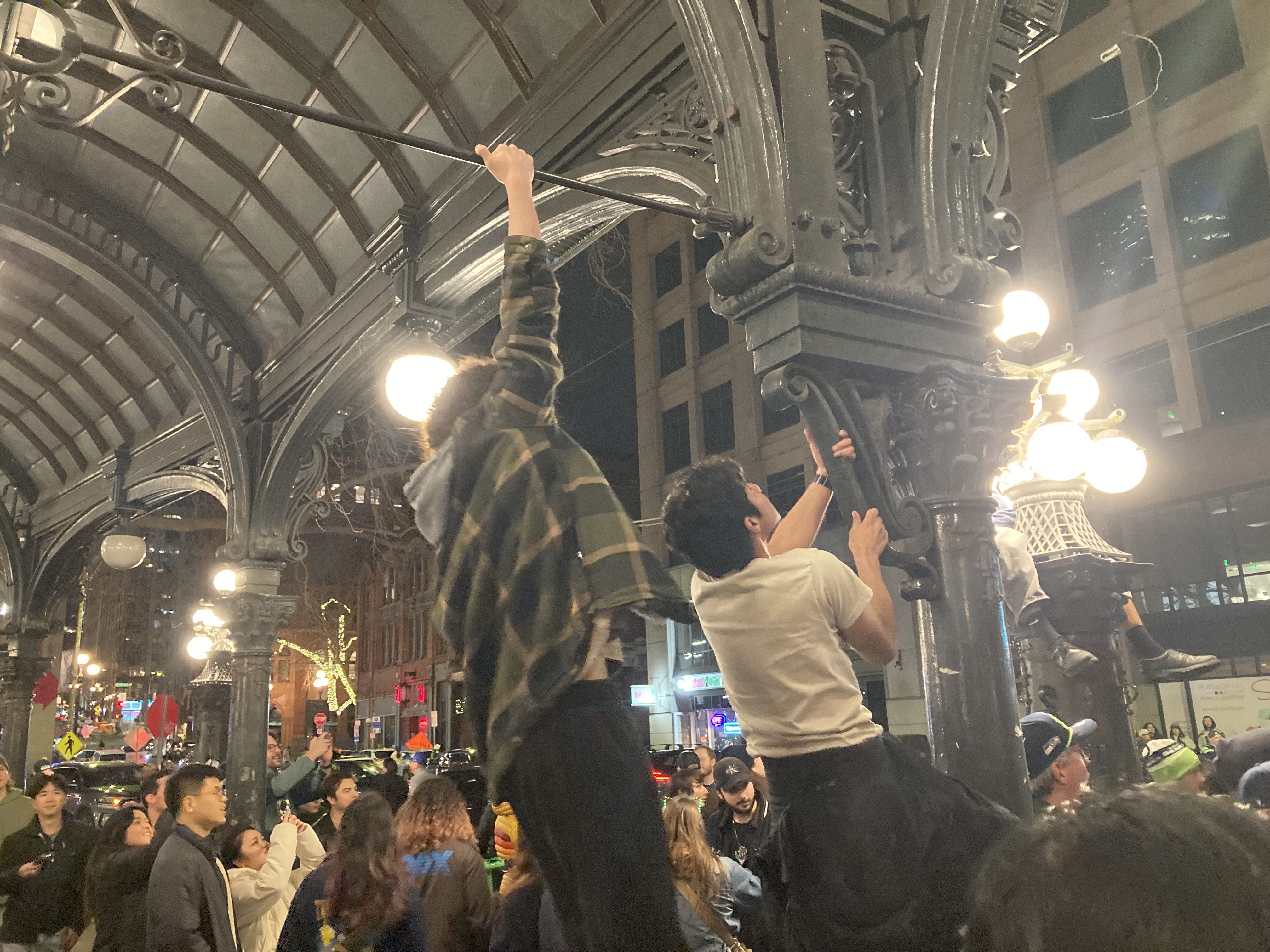 Crowd gathered under a large outdoor metal canopy with ornate columns and bright street lamps at night in a city area; two men climbing the structure while others watch and take photos.