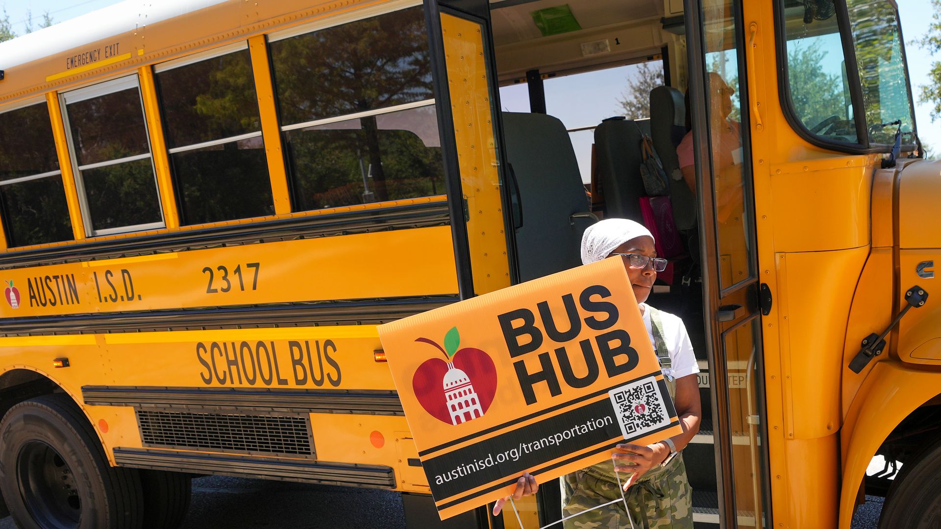 Yellow school bus with an open door; a person wearing a white headscarf and glasses stands by, holding an orange sign that reads "BUS HUB" next to the bus with "SCHOOL BUS" on the side.