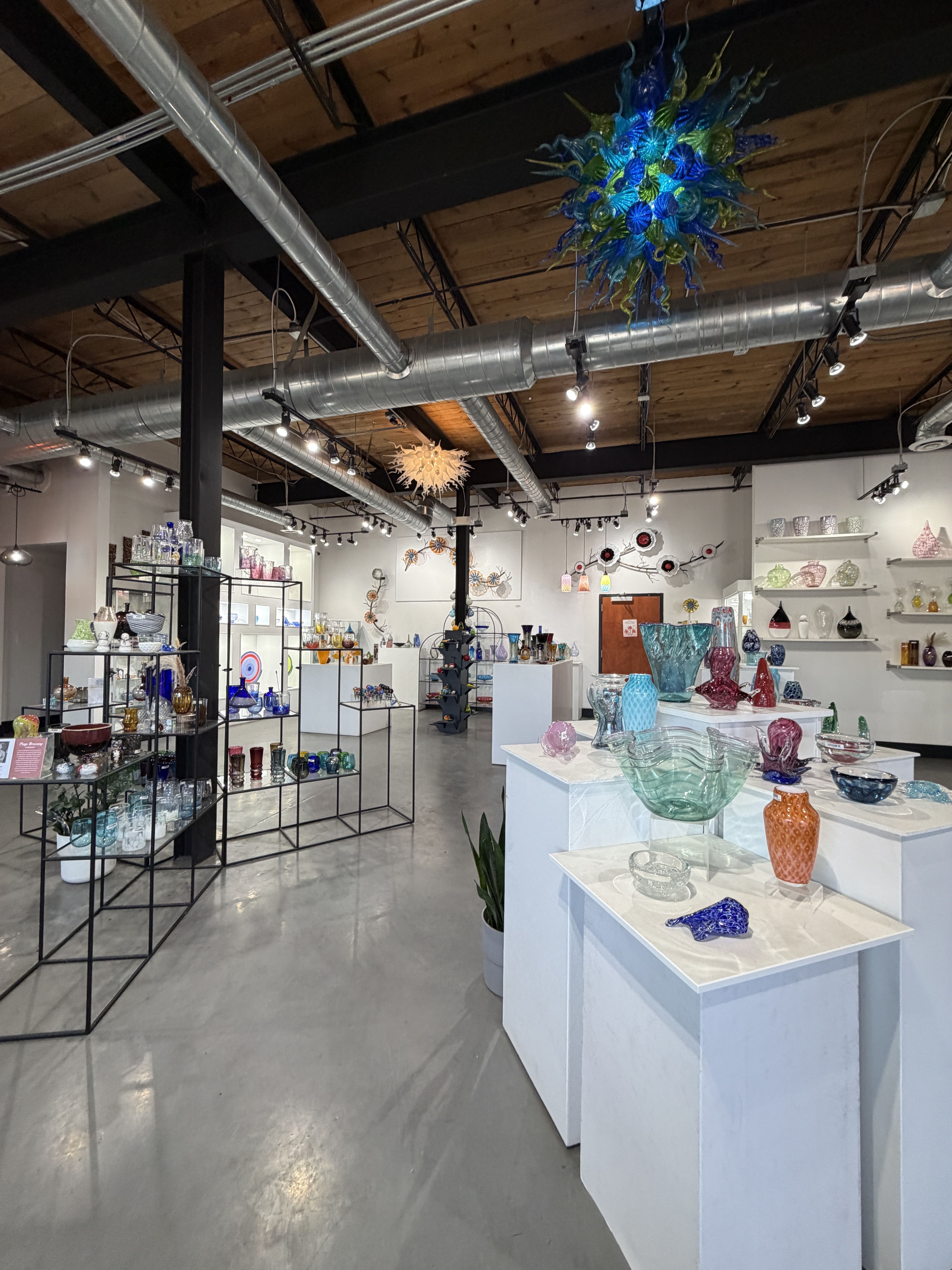 Glass art shop interior with white display pedestals and black metal shelves, colorful glass bowls and vases, and a blue-green glass chandelier hanging from a wooden ceiling with exposed ducts.