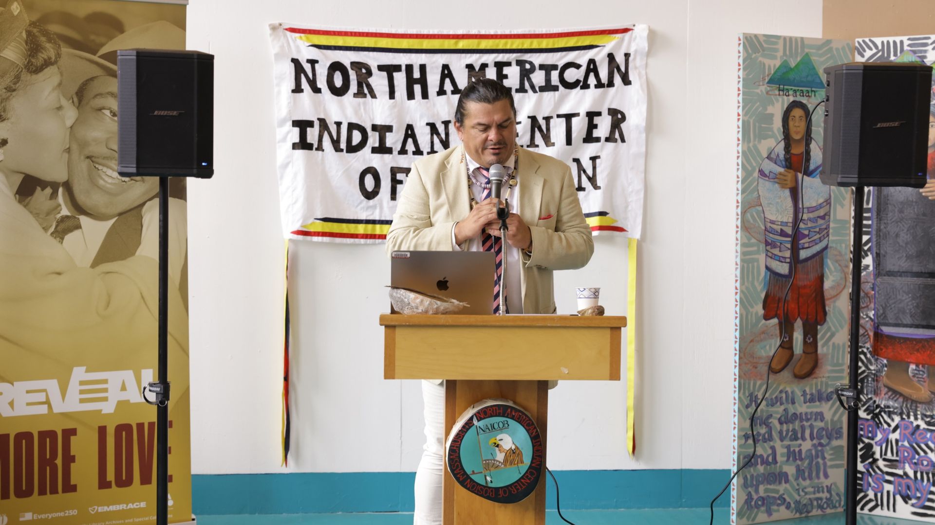 Jean-Luc Pierite, board president of the North American Indian Center of Boston, holds a mic and speaks at a lectern in June 2026 before the unveiling of a marker commemorating NAICOB's legacy.