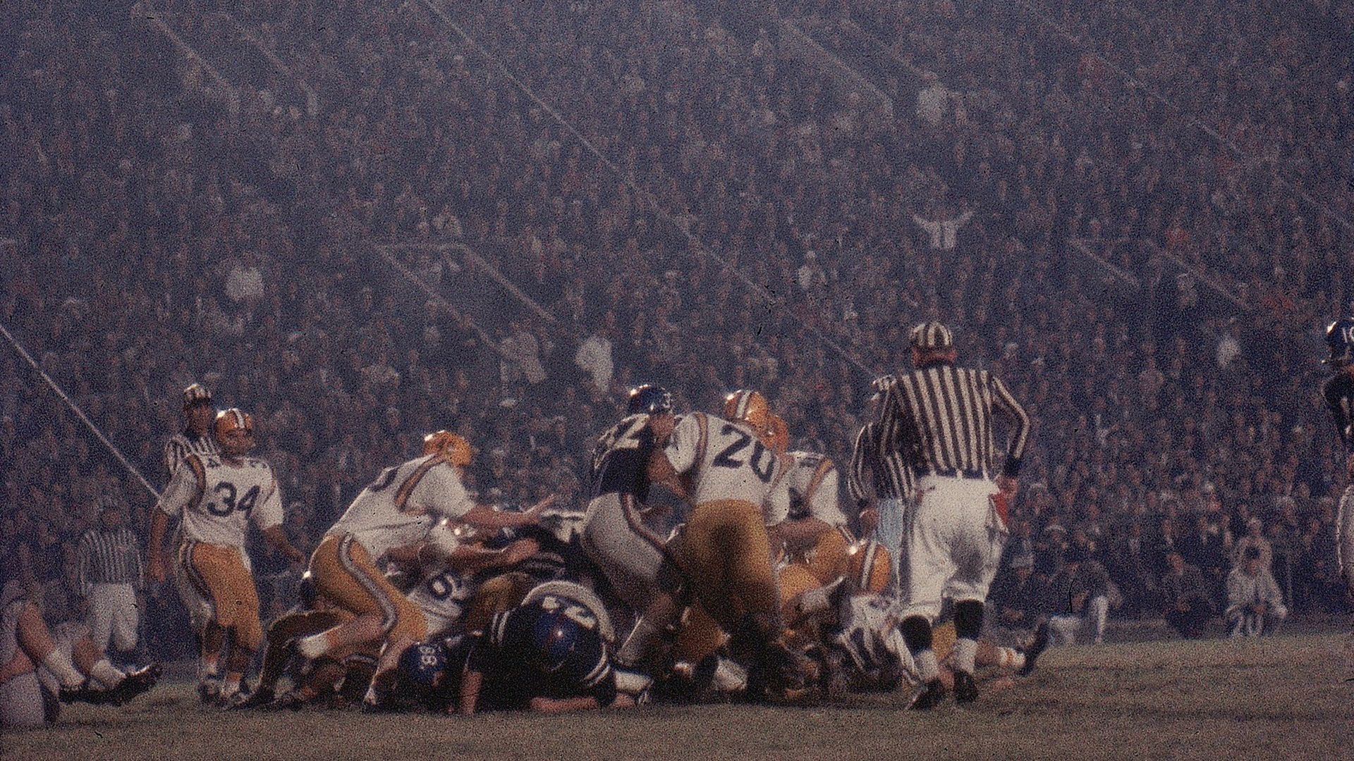 Nighttime football game with players in dark and white-yellow uniforms tackling on field, referees in black-and-white stripes, large crowd in stadium under bright lights.