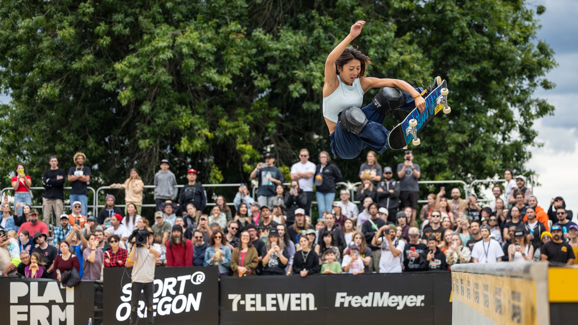 Female skateboarder in light blue top and black knee pads performs a high air trick above a skate ramp, with a large crowd watching behind a black barrier with sponsor logos.
