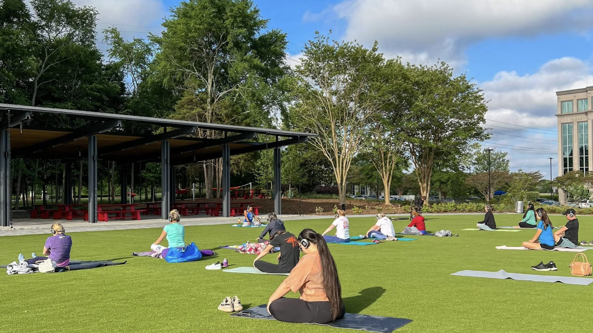 Group of people sitting on yoga mats spaced out on green grass, wearing headphones, under blue sky with white clouds near trees and a pavilion with red benches.