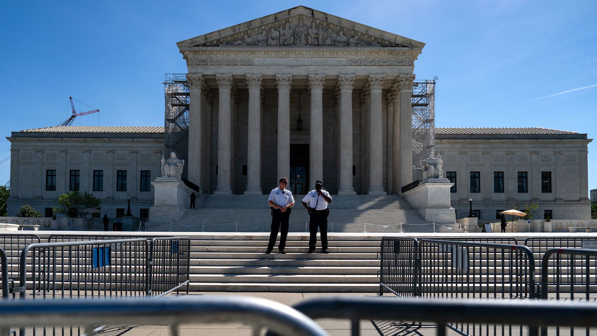 Two officers stand in front of the Supreme Court, a building with columns in the front. The sky is blue in the background. Some metal gates are in the foreground.