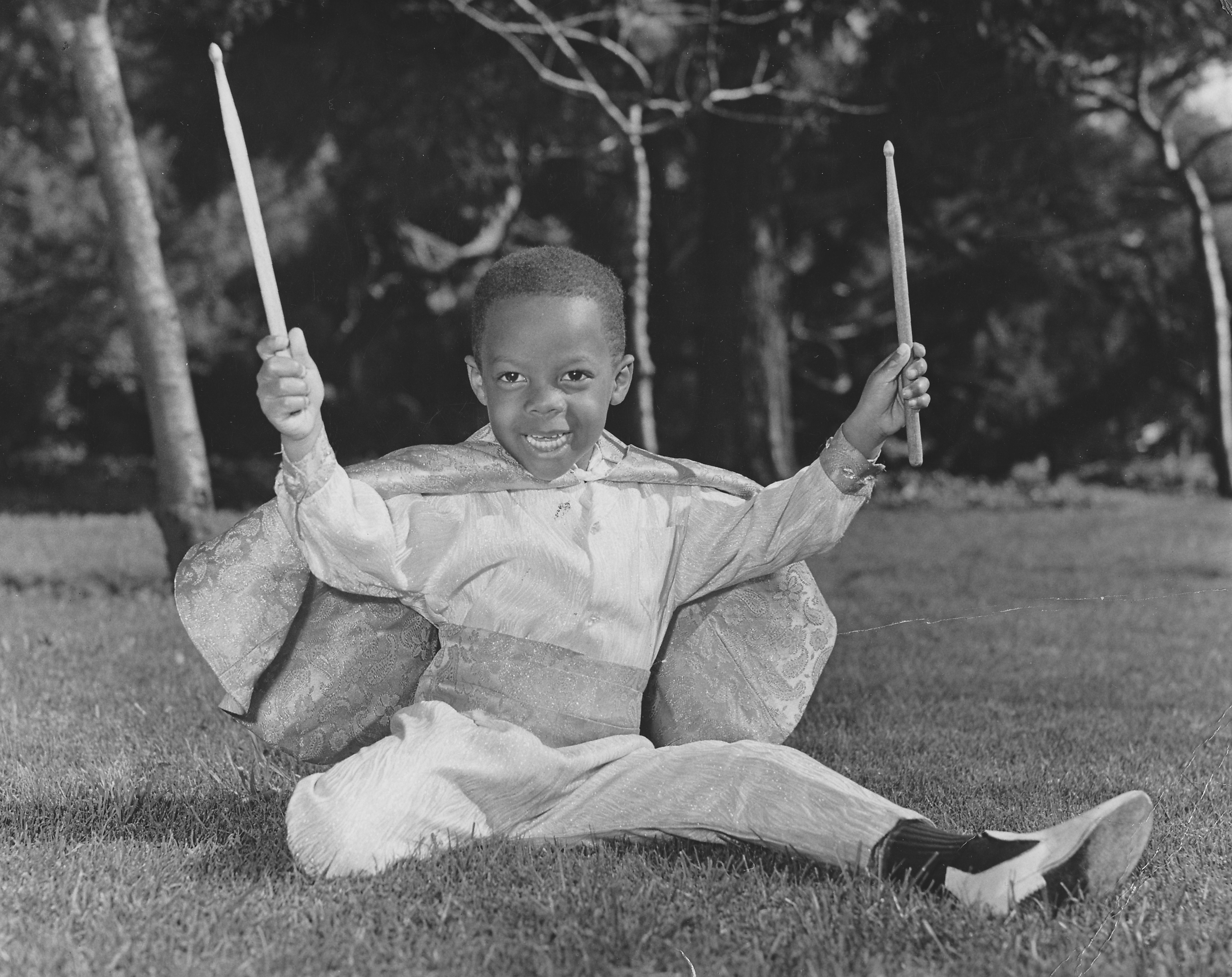 African-American boy sitting in a park, wearing a cape and holding drumsticks.