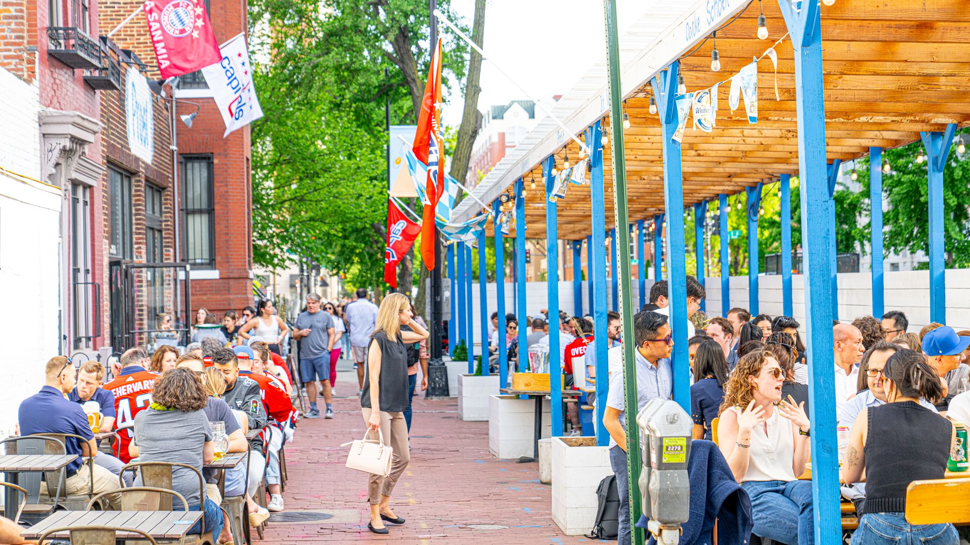 Busy outdoor street festival with a blue wooden pergola on the right, string lights, flags and banners along brick buildings; people sit at tables and stroll beneath green trees.