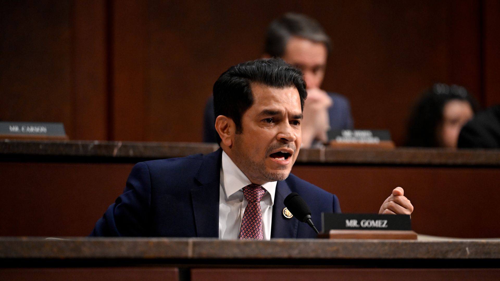 Man in a navy suit and red tie speaks into a microphone at a formal hearing, gesturing with his hand, with blurred officials and a visible nameplate reading 'MR. GOMEZ' in the background.